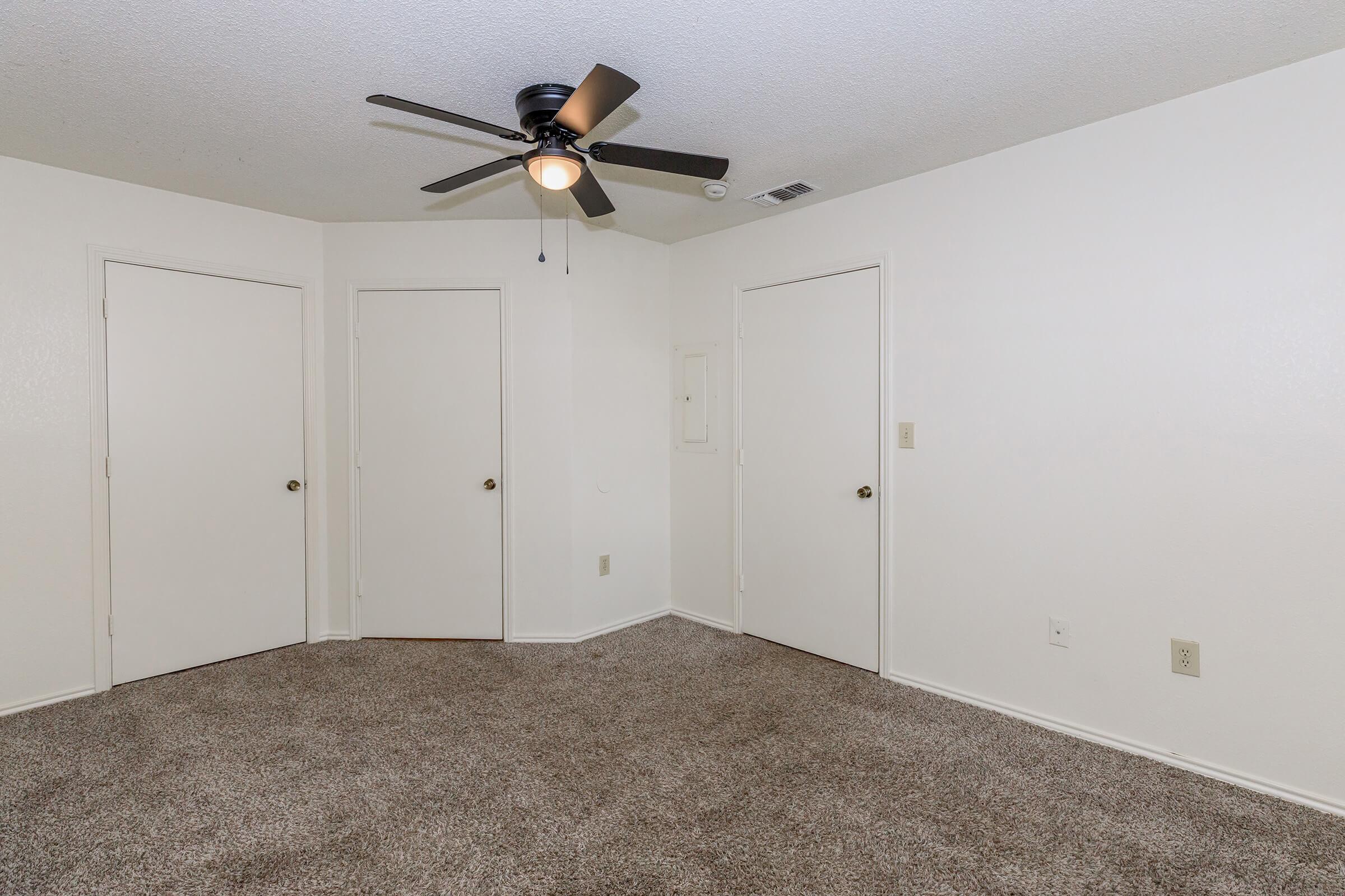 A spacious, empty room with beige carpet flooring. There are three closed white doors along two walls, and a ceiling fan with a light fixture is mounted in the center. The walls are painted white, creating a bright and open atmosphere.