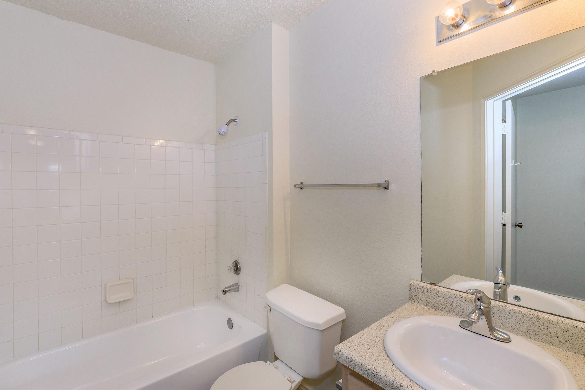 A clean, simple bathroom featuring a white tiled bathtub with a showerhead, a toilet, and a sink with a granite countertop. There is a wall-mounted light fixture above the mirror, and a doorway is visible in the background, leading to another room. The walls are a light color, creating a bright and airy atmosphere.