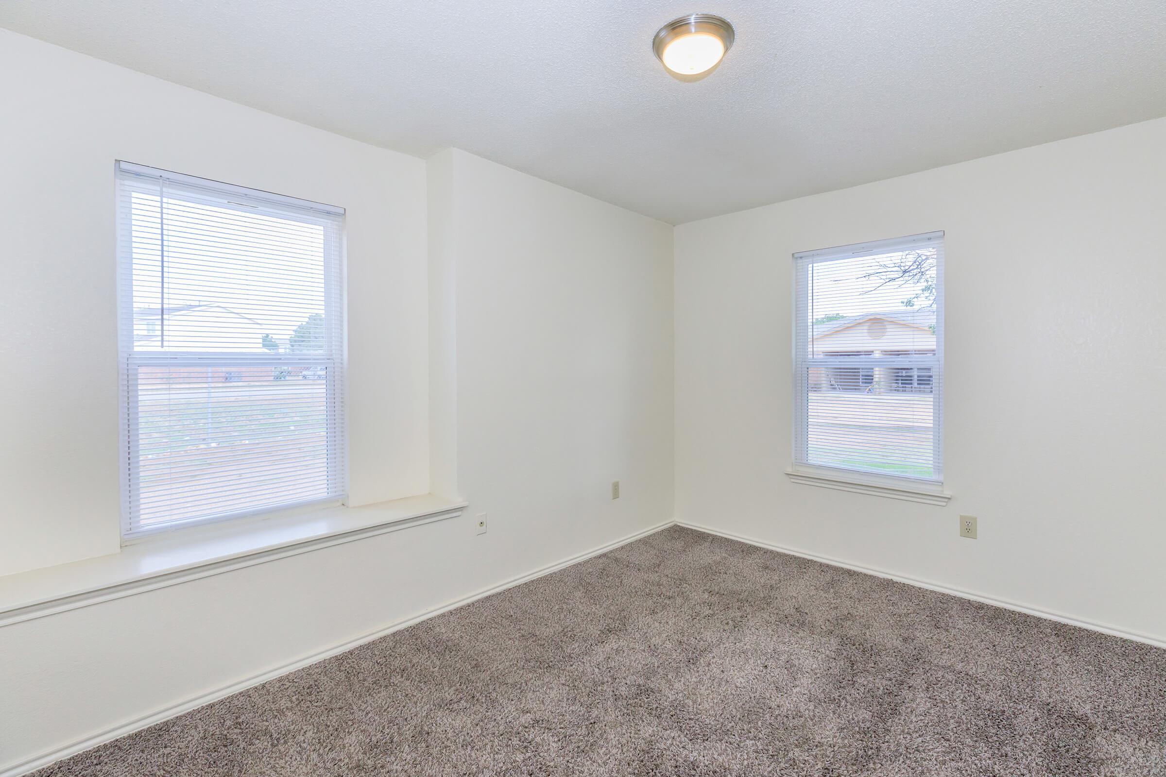 A bright, empty room with beige carpet and white walls, featuring two windows with blinds letting in natural light. The room has a ceiling light fixture and minimal furnishings, creating a clean and spacious appearance.