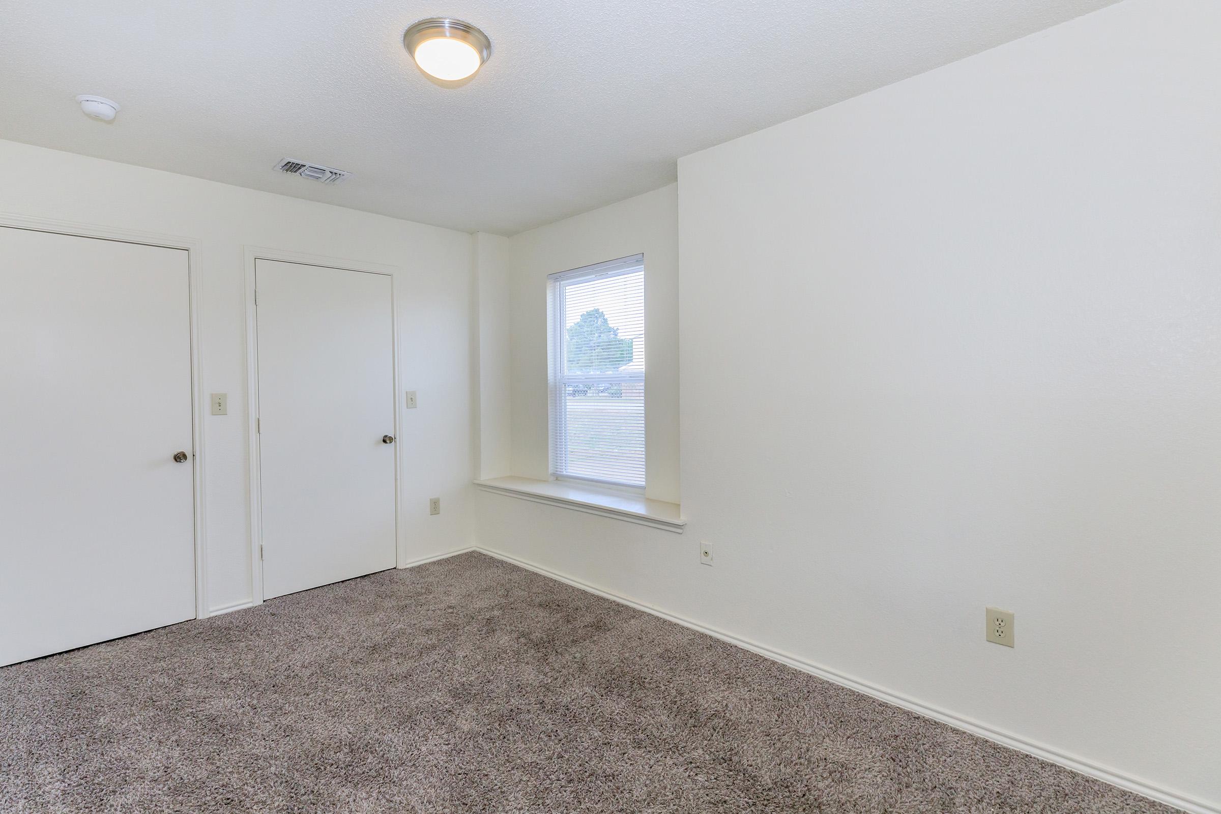 A spacious, empty room with light gray carpet, a single window allowing natural light, and a white wall. There are two doors: one leading to a closet and another possibly to another space. The ceiling features a modern light fixture. The overall atmosphere is bright and inviting.