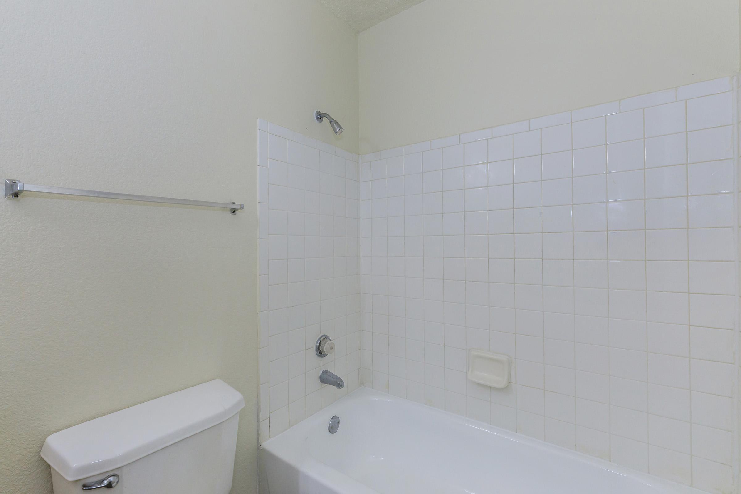 A clean, simple bathroom featuring a bathtub with showerhead, a white tiled wall, and a toilet. The walls are painted a light color, and there is a towel rack on one side. The space is well-lit and uncluttered, creating a serene atmosphere.