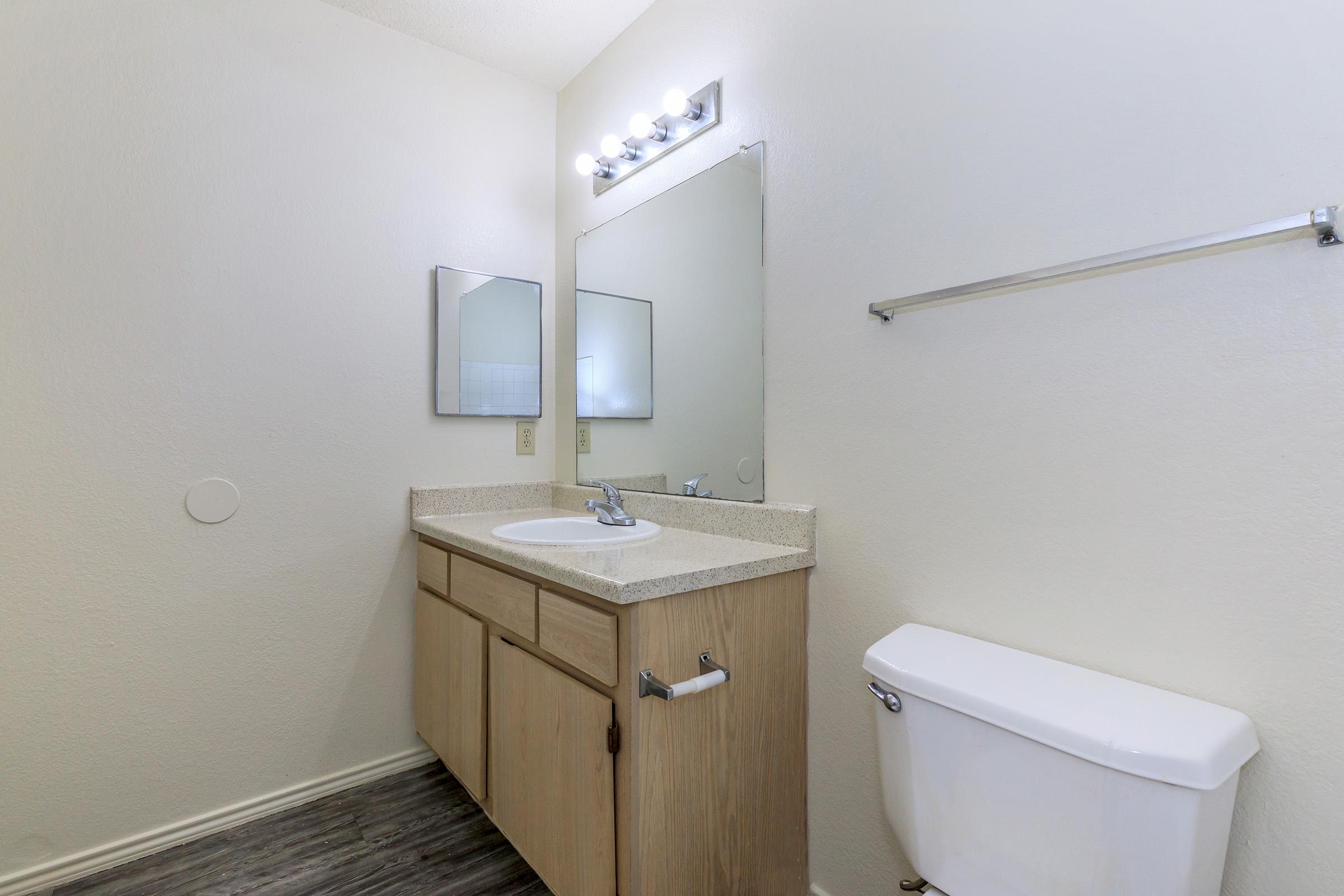 A clean and simple bathroom featuring a wooden vanity with a sink, a large mirror above, a wall-mounted light fixture, a toilet, and light-colored walls. The flooring is dark and contrasts with the overall light tone of the space.