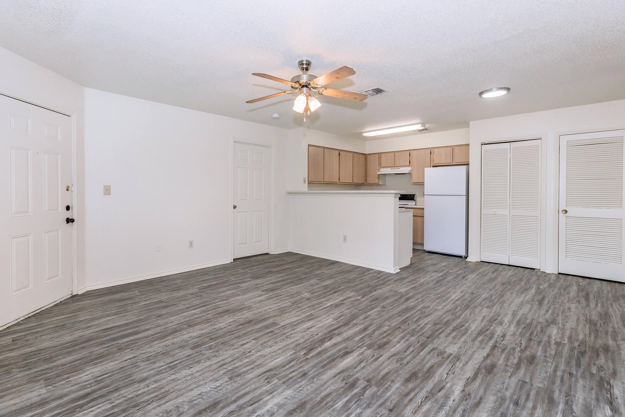 A spacious, minimalist living area with light wood-look flooring, a ceiling fan, and a white kitchen featuring beige cabinets. The room has a large window, a white refrigerator, and two doors leading to other areas. The walls are painted white, creating a bright, airy atmosphere.