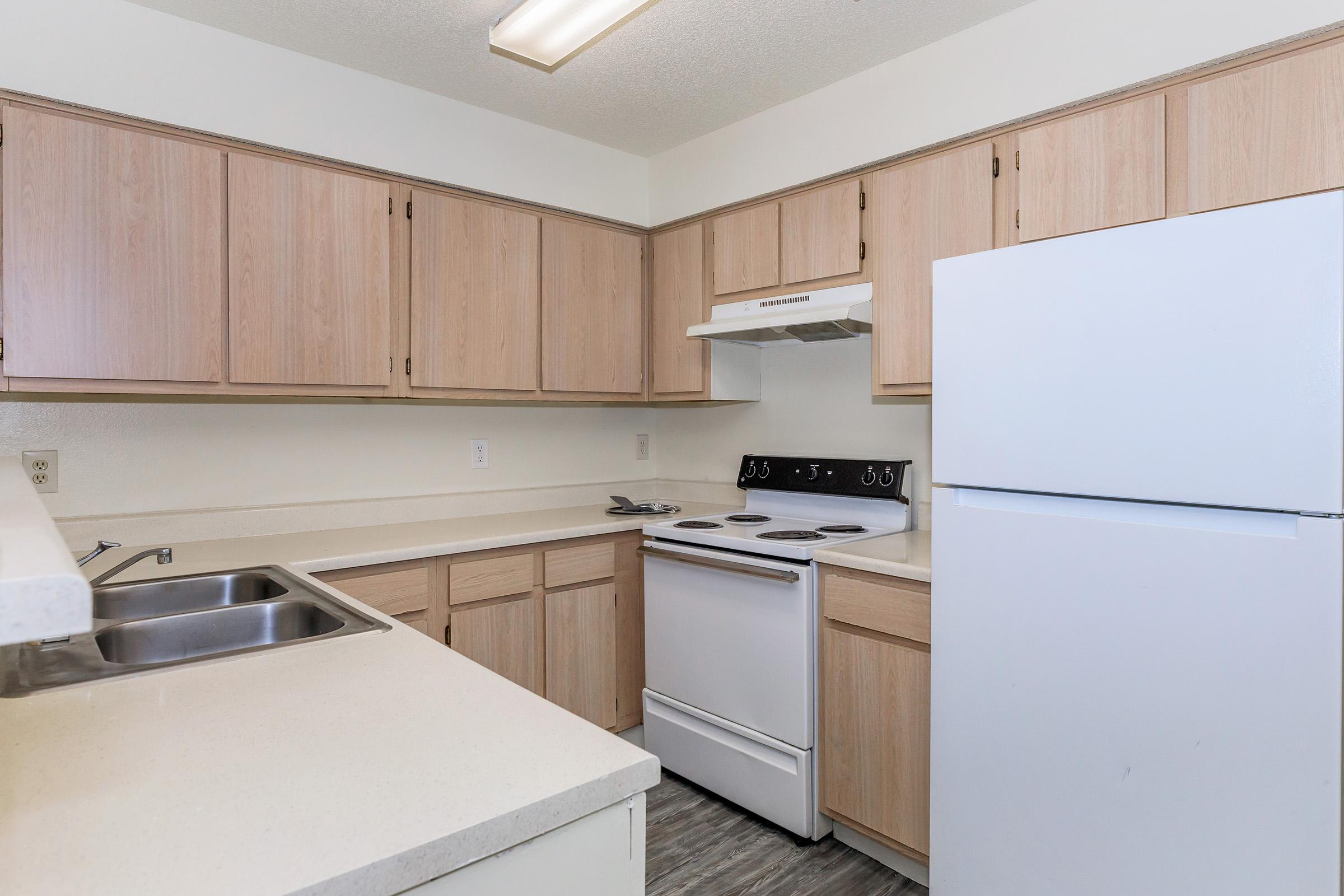 A compact kitchen featuring light wooden cabinetry, a white refrigerator, a white stove with an oven, and a double sink. The countertops are light-colored, and the flooring appears to be a soft gray. The kitchen is well-lit with overhead lighting, creating a clean and inviting space.