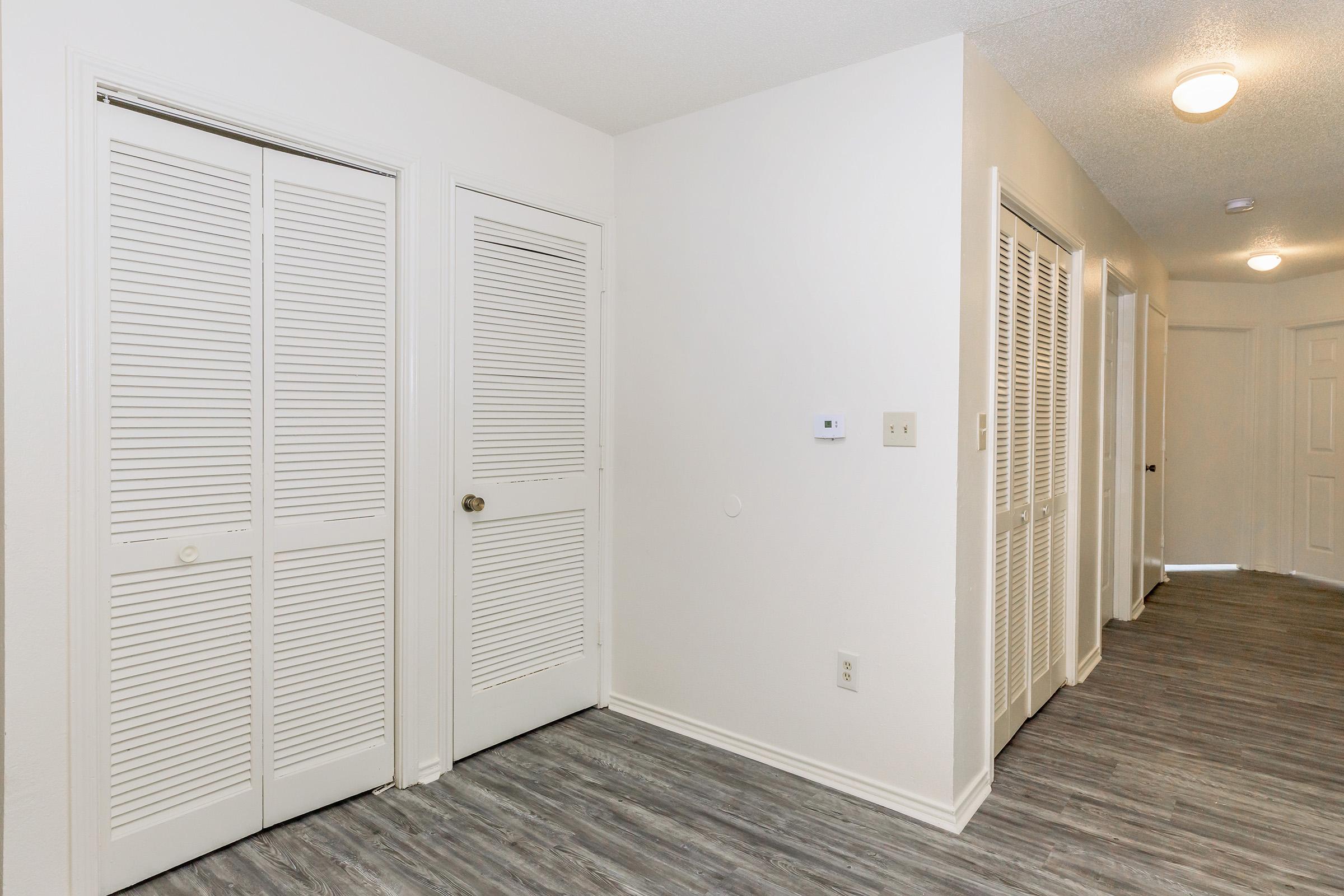 A well-lit hallway featuring white walls and gray wood-like flooring. Two doors with white louvered panels are visible on the left, and additional doors are seen along the right side. A thermostat is mounted on the wall near the left doors, adding to the modern and clean appearance of the space.