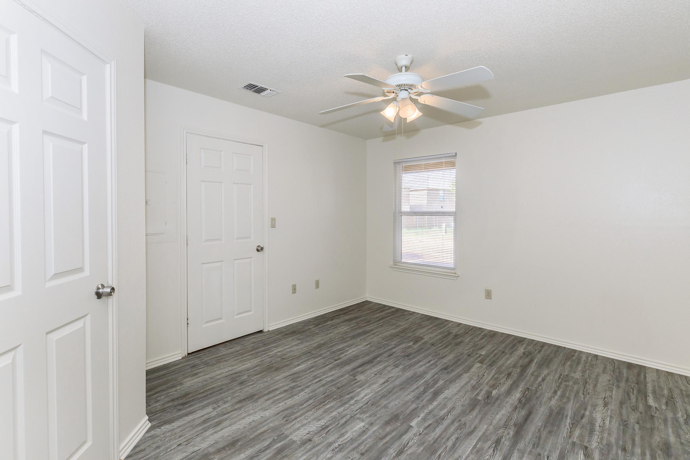 A bright, empty room with light-colored walls and a ceiling fan. The floor features wood-like laminate, and there are two doors: one leading to a closet and another to the exterior. A window allows natural light to enter, creating a spacious and airy feel.