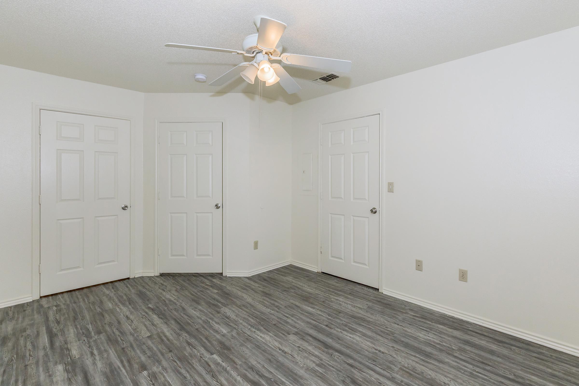 A spacious, well-lit room featuring light-colored walls, a ceiling fan, and a modern laminate floor. The room includes two closed doors, likely leading to closets or other areas, and has a neutral aesthetic suitable for various decor styles.