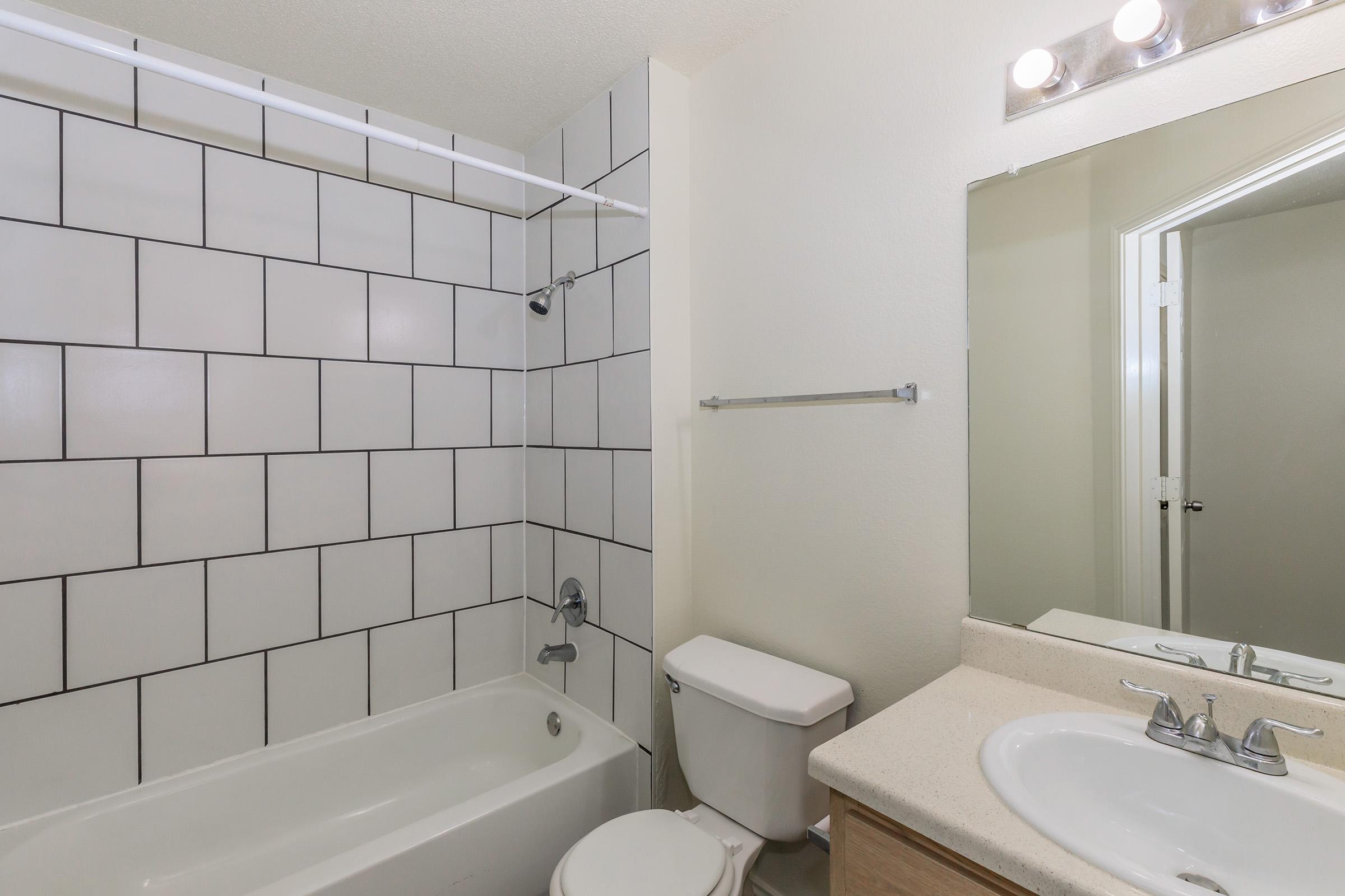 A modern bathroom featuring a bathtub with a showerhead, white tiled walls with dark grout, a toilet, and a vanity with dual sinks and a large mirror. The space is bright with neutral colors, and there is a towel rack on the wall. A doorway can be seen leading to another room.