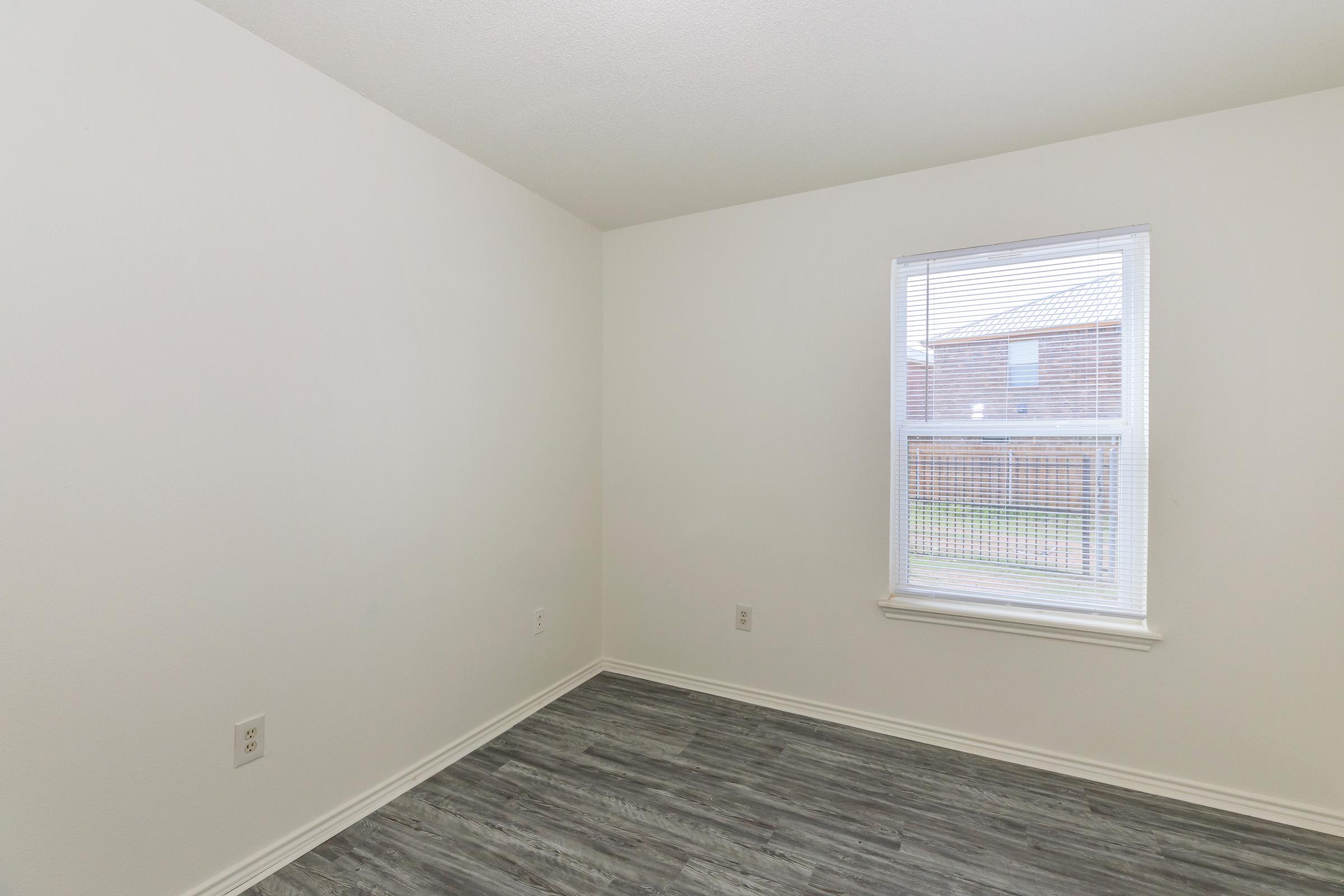 Empty room with light-colored walls and a window letting in natural light. The floor features modern gray laminate flooring, and the window has white blinds. The space appears clean and minimalistic, suitable for various uses.