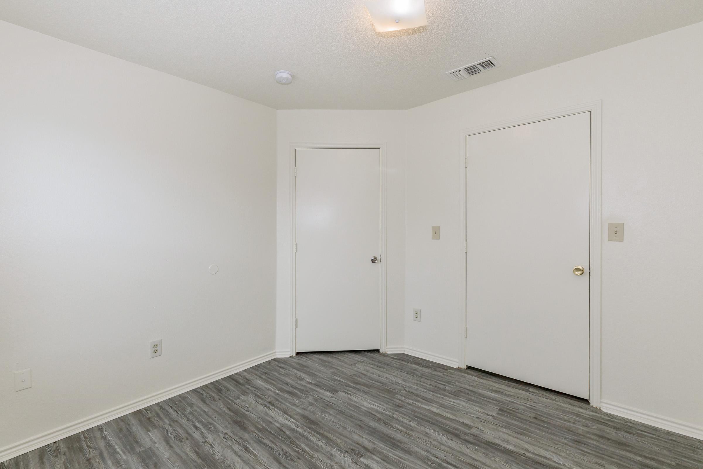 A well-lit, empty room with light-colored walls and a laminate floor. There are two closed doors on opposite walls, and minimal furniture or decor, creating a clean and simple appearance.
