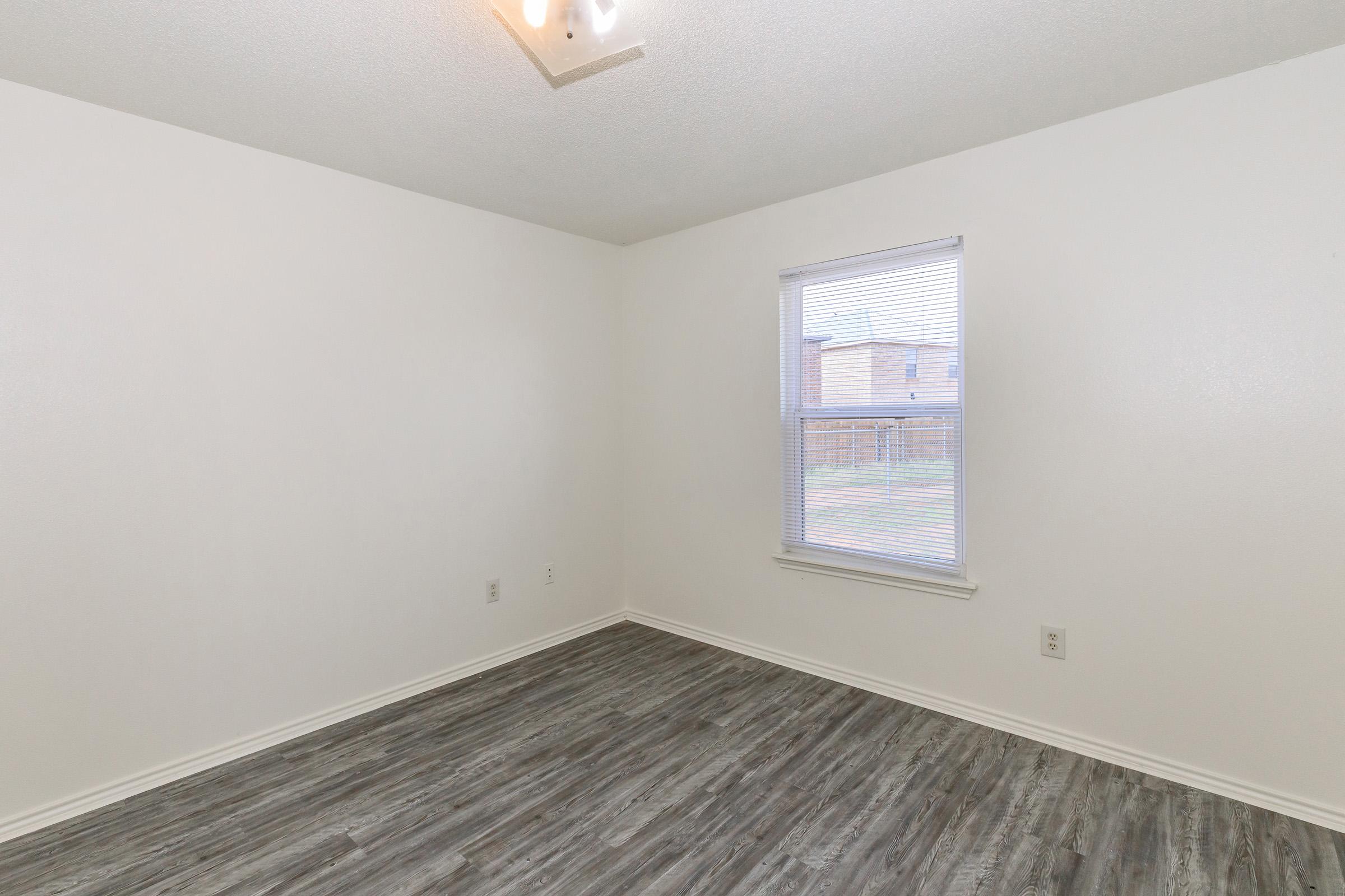 Empty room with light-colored walls and a large window allowing natural light. The floor features a modern, gray wooden texture. A ceiling light fixture is visible above, and there are no furnishings in the space, providing a clean and minimalistic appearance.
