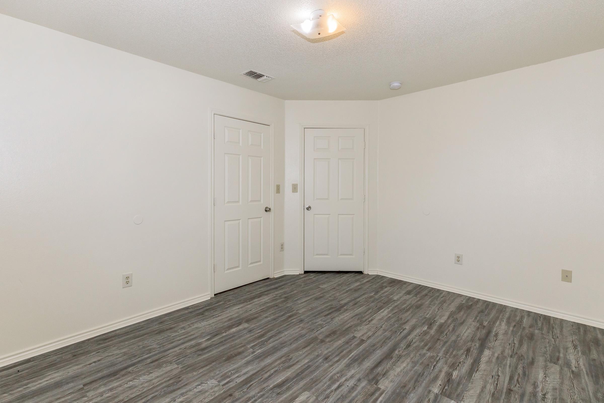 A clean, empty room with light-colored walls and a textured, gray floor. There are two closed doors on the left side, one directly in front, and a ceiling light fixture illuminating the space. The room lacks furniture, providing a simple and spacious feel.