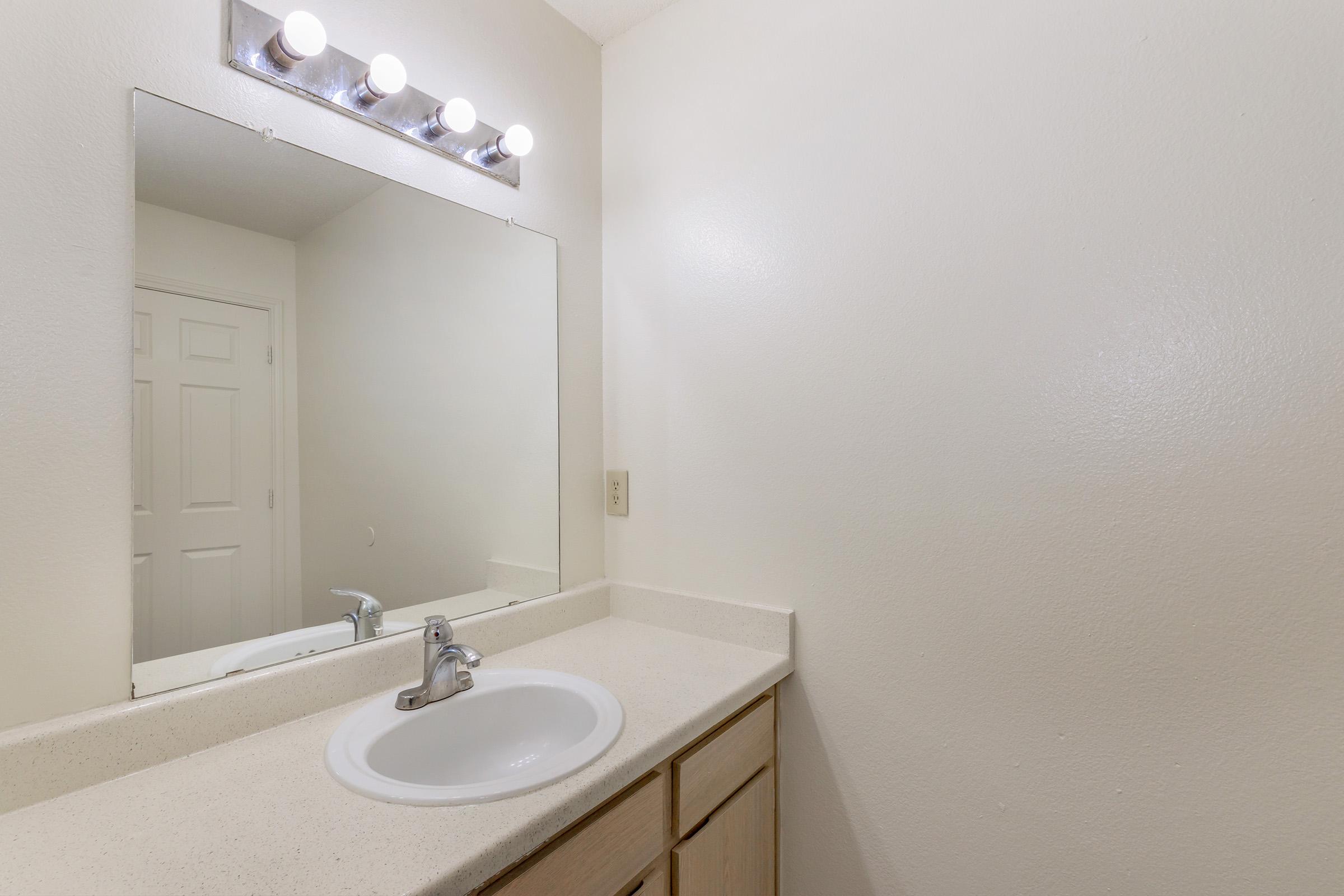 A clean, simple bathroom featuring a white countertop with a sink, a large mirror above the sink, and modern light fixtures. The walls are a light color, and there is a door visible in the background, suggesting a minimalistic design.