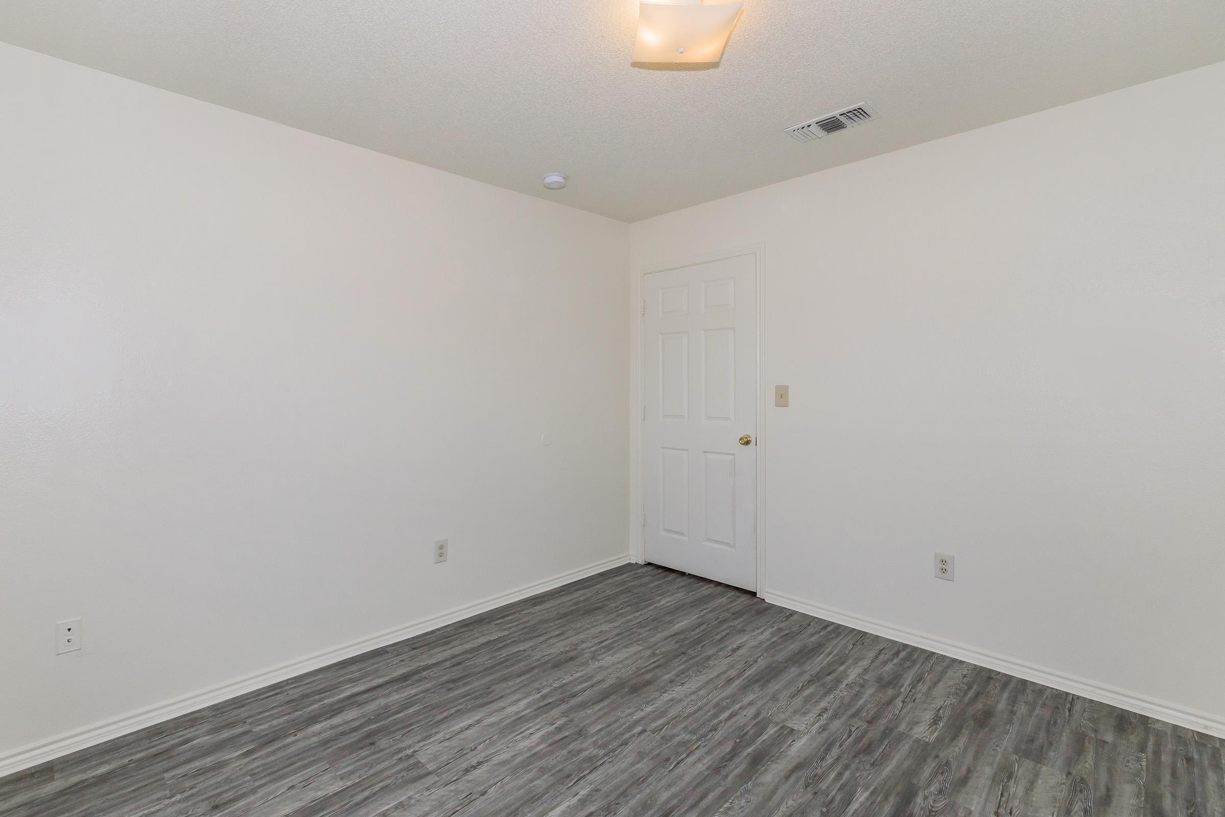Empty room with light gray flooring and white walls. A single light fixture in the ceiling illuminates the space. There is a closed white door on one wall, and no furniture or decorations are present, creating a minimalistic atmosphere.