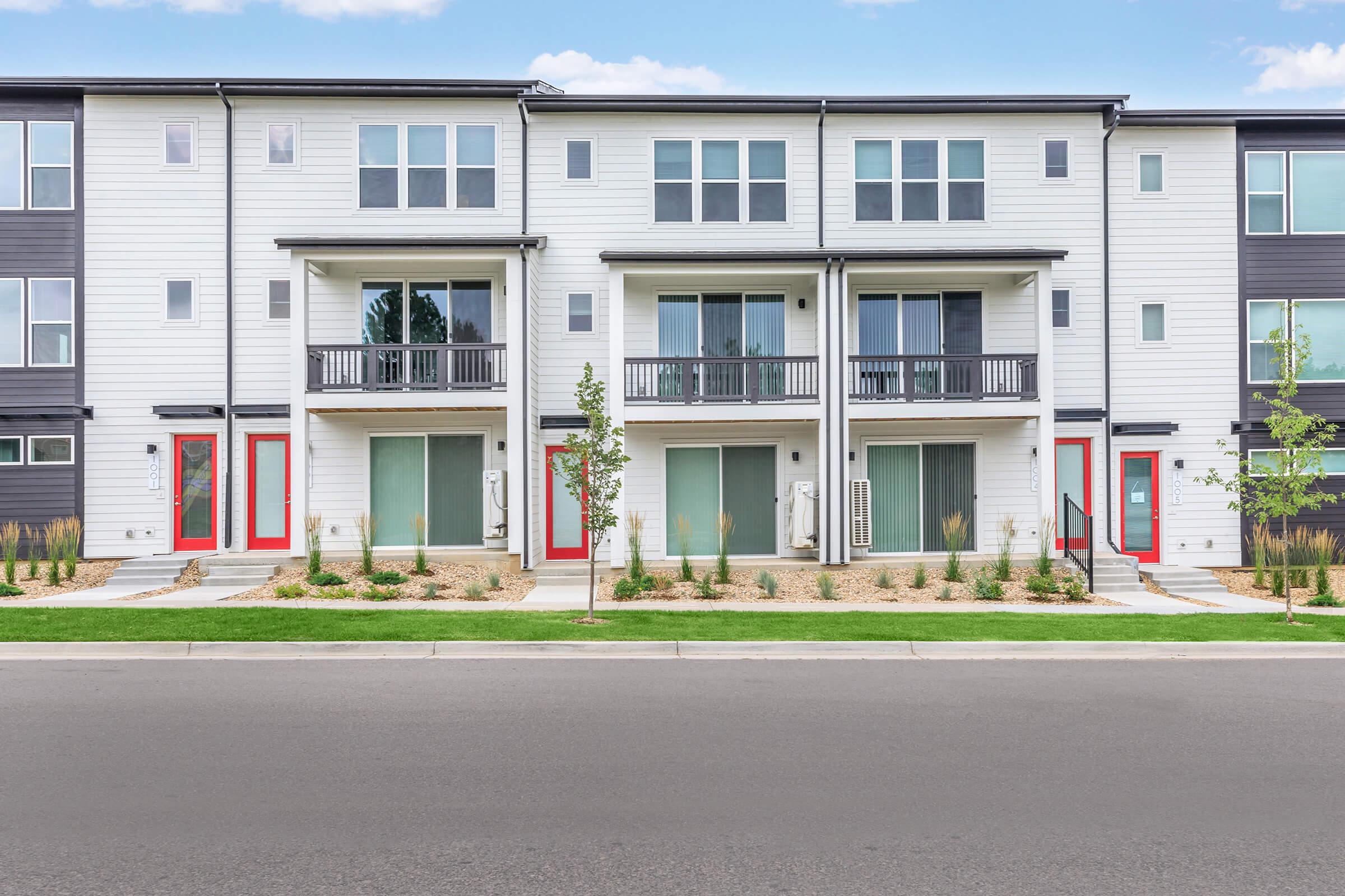 A modern multi-unit residential building featuring two stories with a mix of white and dark exterior. Each unit has a balcony, large windows, and red front doors. The landscape includes small shrubs and grass in front of the building along a paved road.