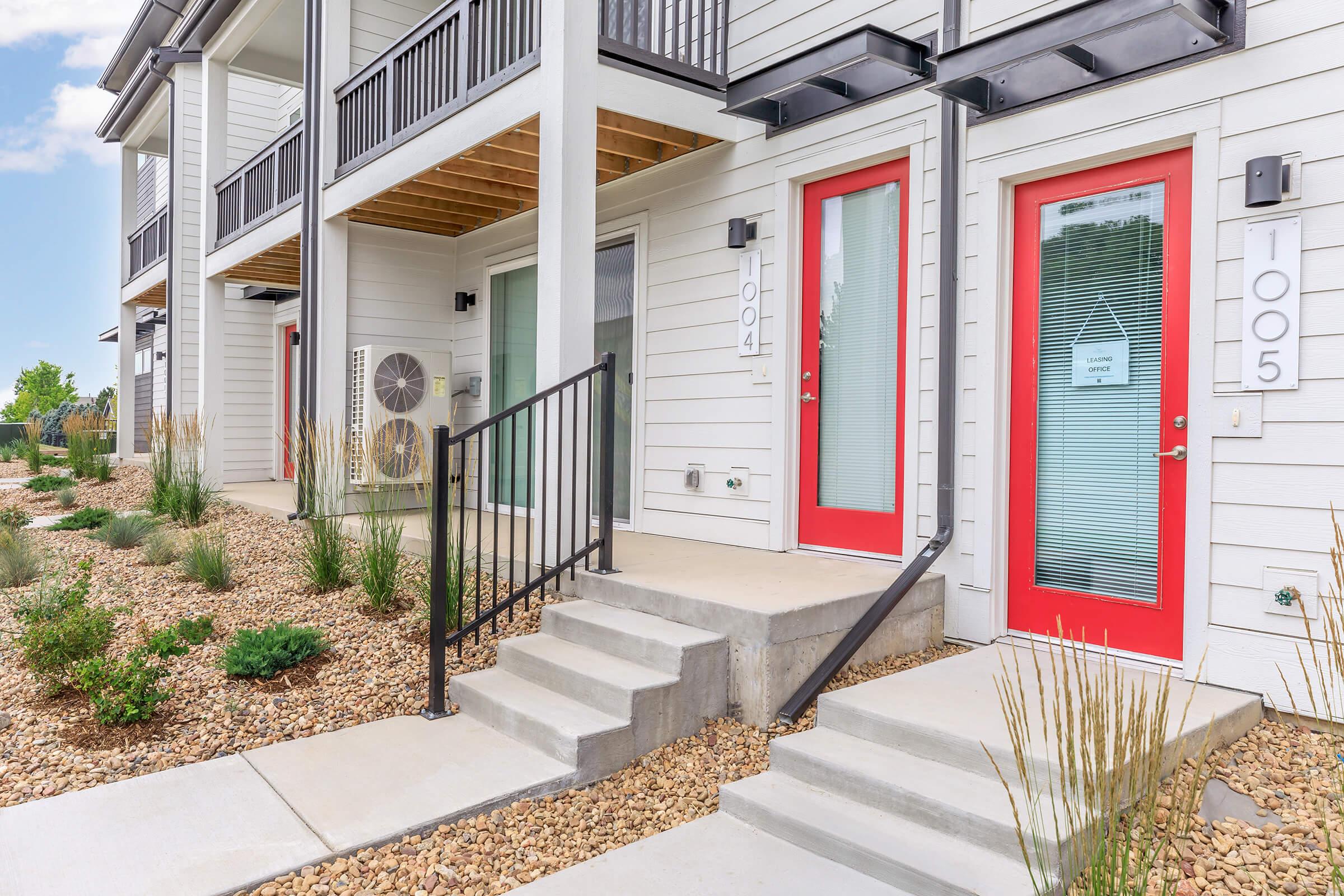 Two adjacent modern townhome entrances featuring red doors, with visible unit numbers 1004 and 1005. There is a small staircase leading to each door, surrounded by a landscaped area with rocks and sparse greenery. Clear blue skies and a well-maintained exterior emphasize a contemporary residential aesthetic.