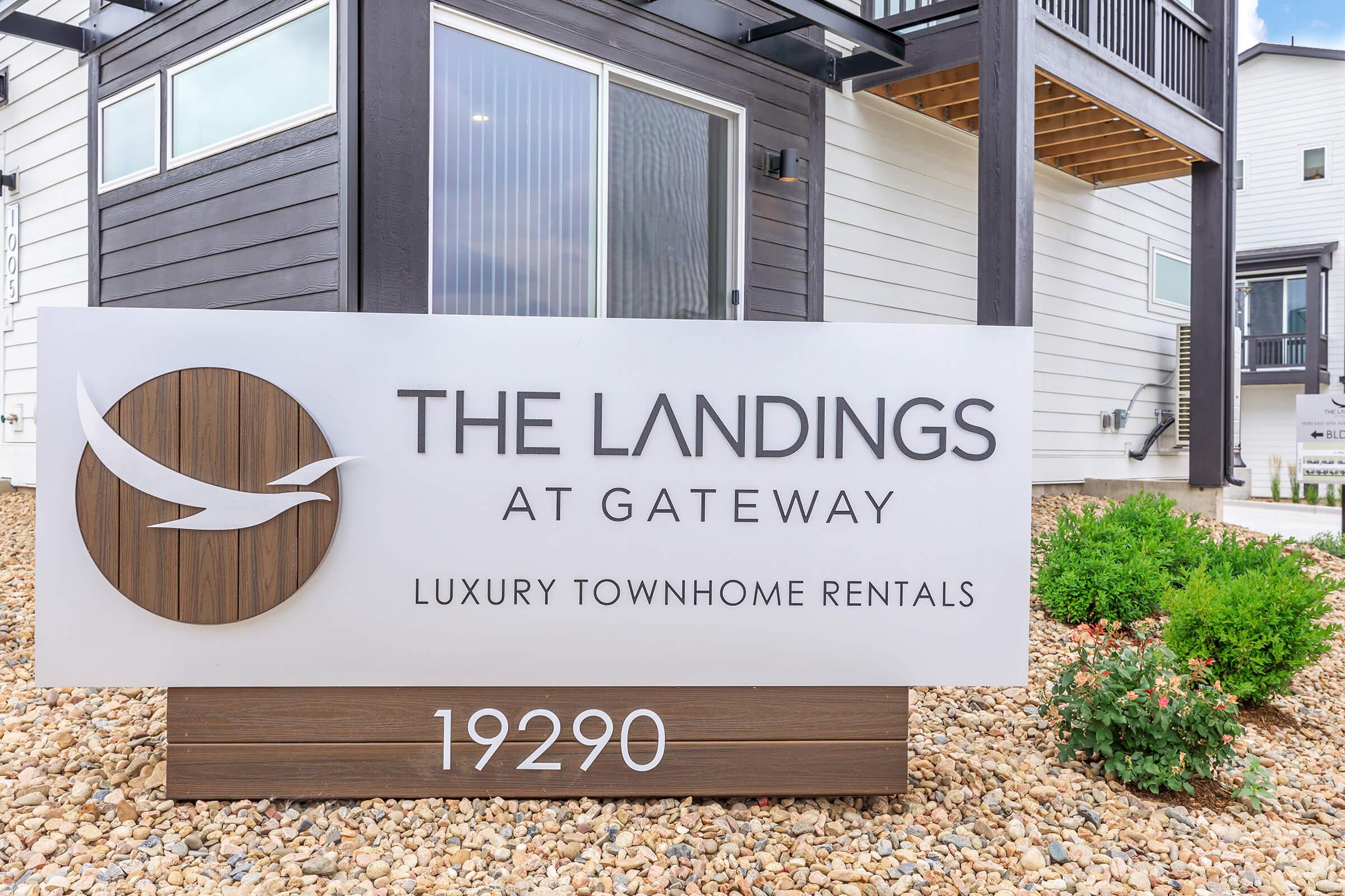 Sign for "The Landings at Gateway," featuring the name prominently along with the tagline "Luxury Townhome Rentals." The sign is positioned in front of modern townhome buildings with a landscaped area of small shrubs and decorative gravel. Visible address: 19290.