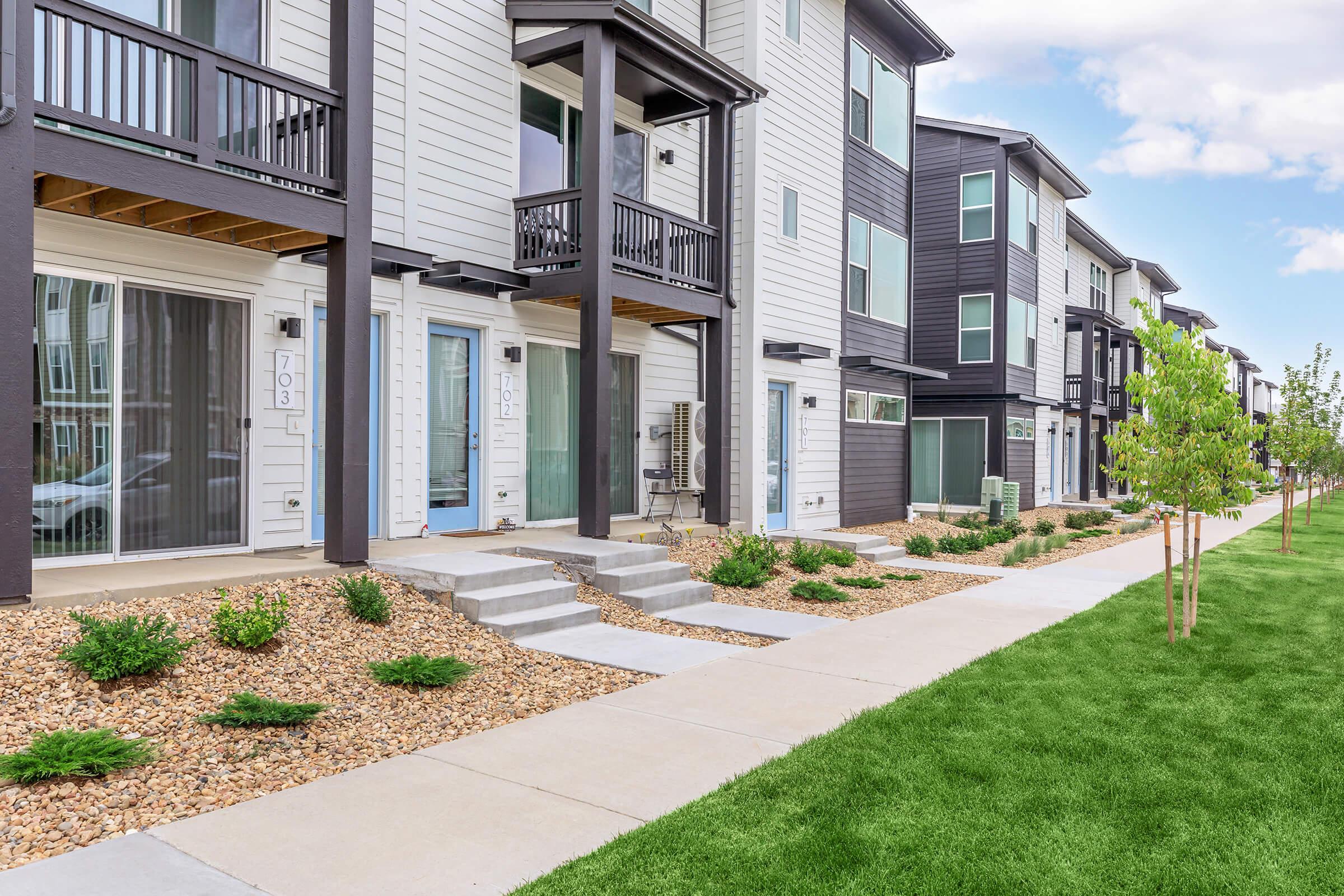 A row of modern townhouses with multiple stories, featuring balconies, large windows, and a nicely landscaped pathway. The front yards have decorative rocks and small trees, while a green lawn stretches alongside the walkway. The buildings have a mix of light and dark siding colors.