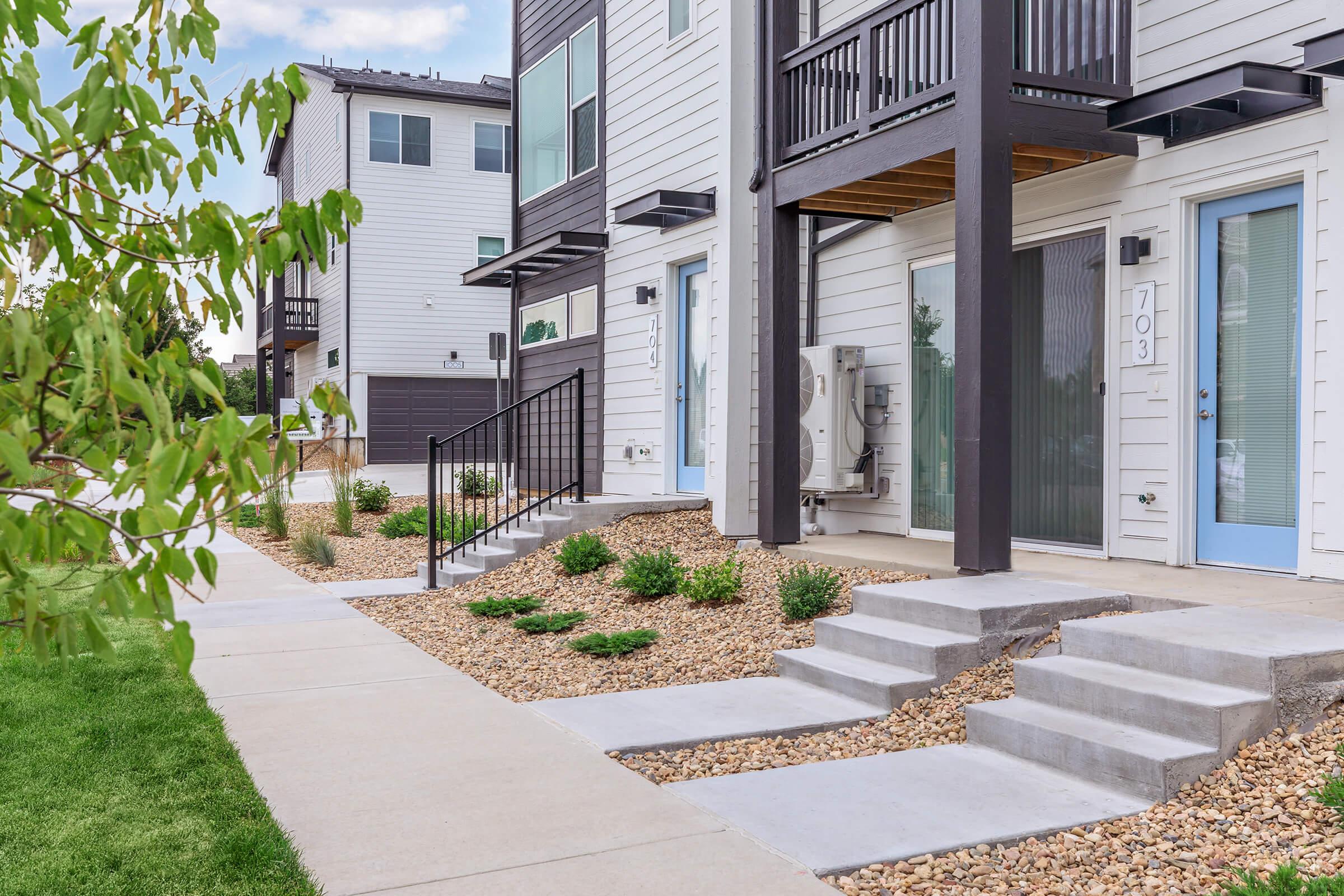 A modern apartment complex featuring two-story buildings with a contemporary design. The exterior showcases light-colored siding, blue doors, and small concrete steps leading to individual units. The landscaping includes gravel and low-maintenance plants, with a well-maintained sidewalk and green lawn.