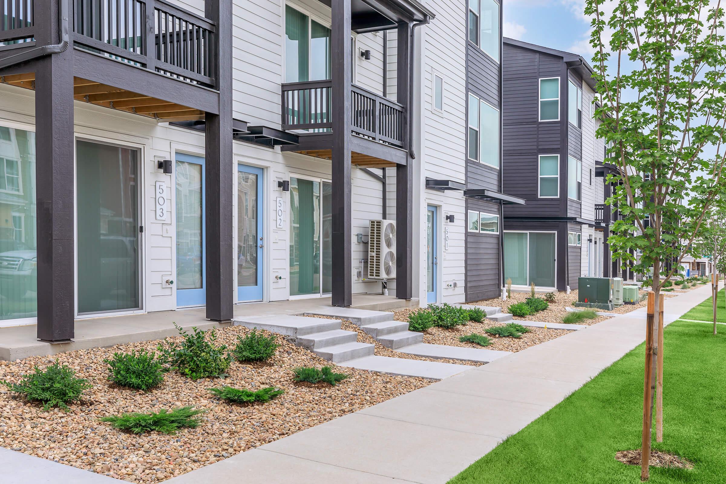 Modern multi-unit residential building with a well-maintained exterior. Features include balconies, stone pathway, and landscaped areas with small shrubs and grass lawns. Utility units can be seen on the side, and street features a concrete walkway. Bright sky above adds to the inviting atmosphere.