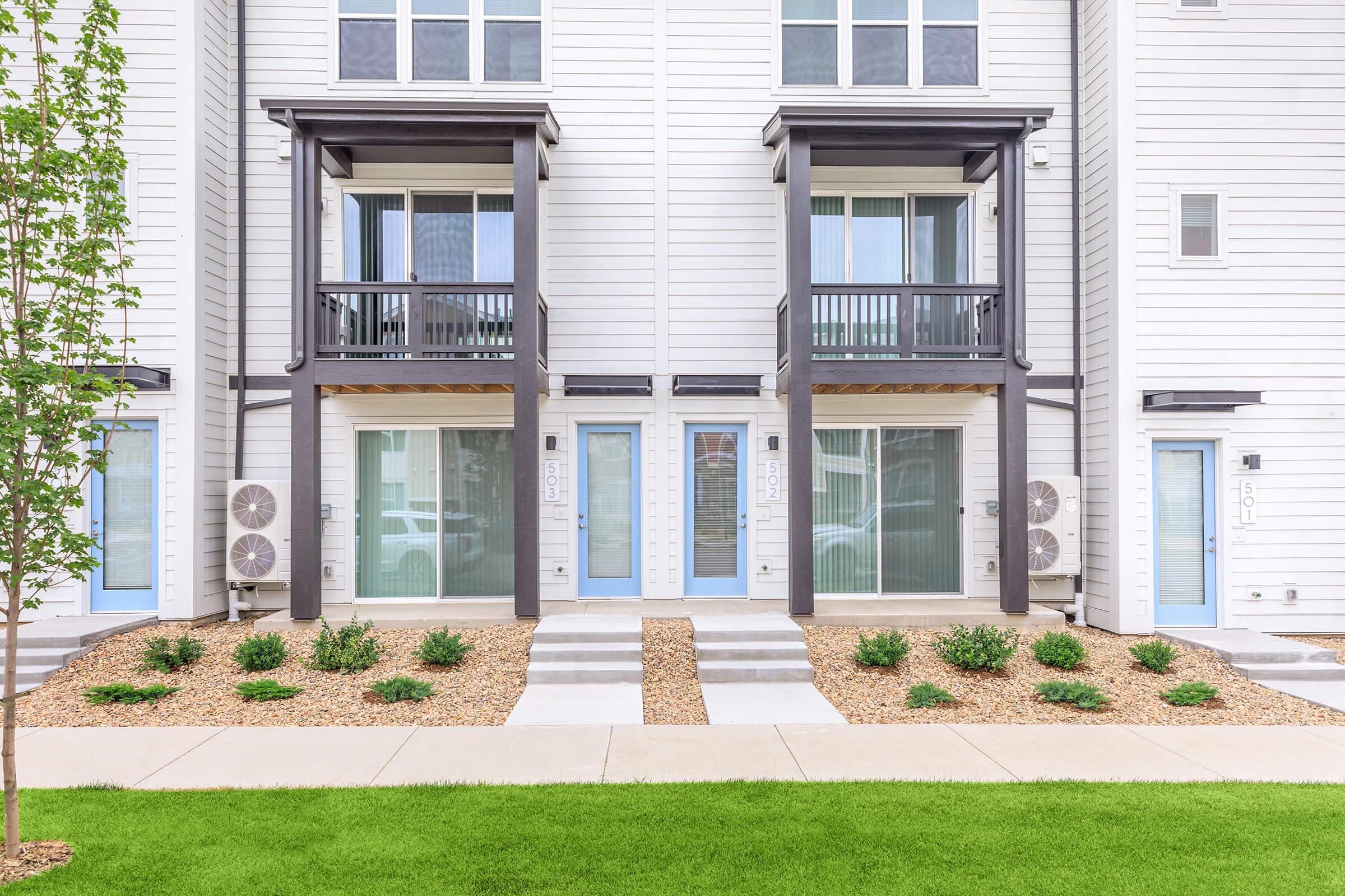 Modern multi-unit residential building with two identical units, each featuring a front porch and large windows. The ground is landscaped with small shrubs and decorative gravel, and there's a manicured green lawn in the foreground. Air conditioning units are mounted outside both units.