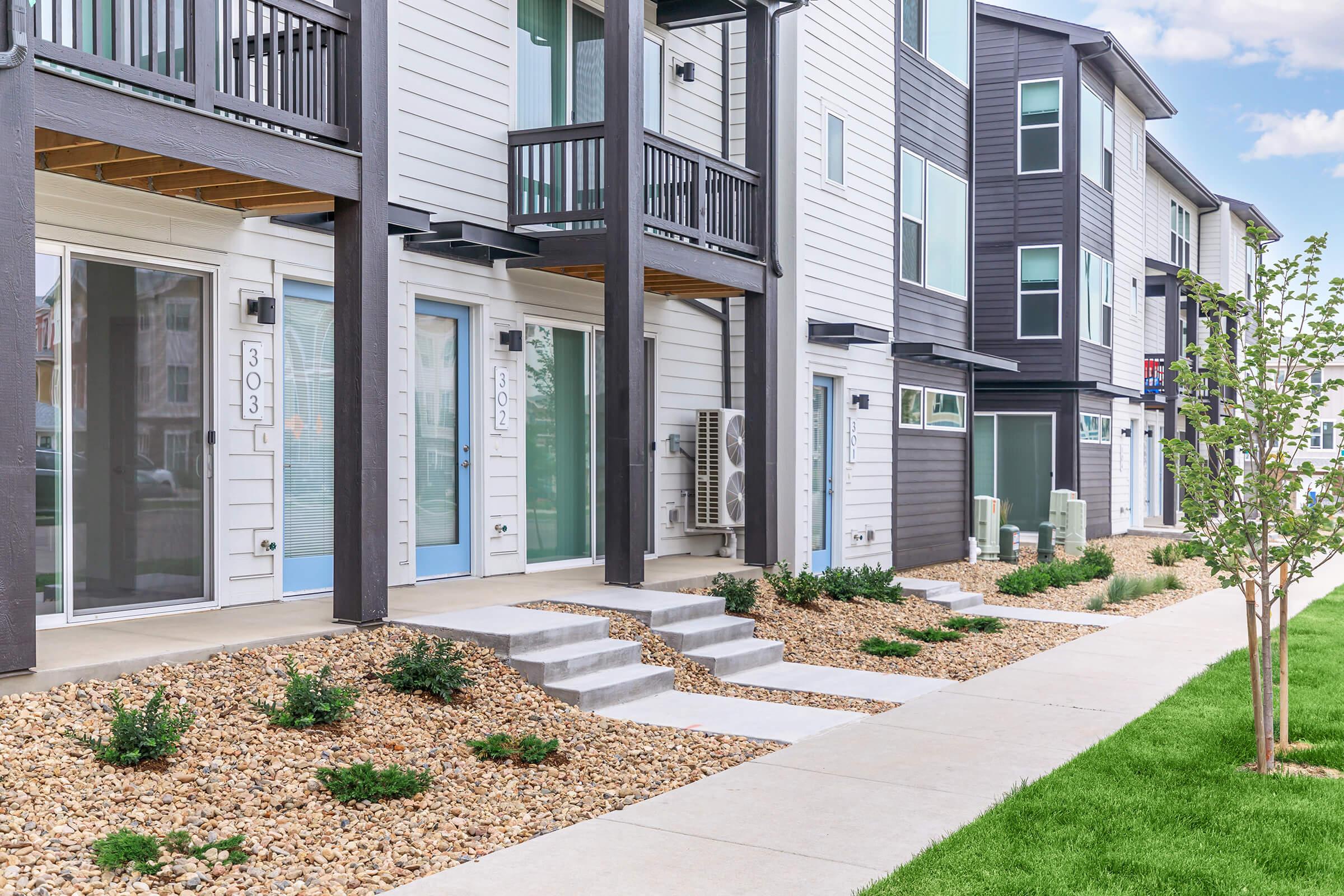 Modern multi-unit residential buildings with balconies and large windows, featuring a clean, landscaped pathway lined with small shrubs and gravel. The structure showcases a mix of white and dark exterior panels, set against a backdrop of green lawn and blue sky.