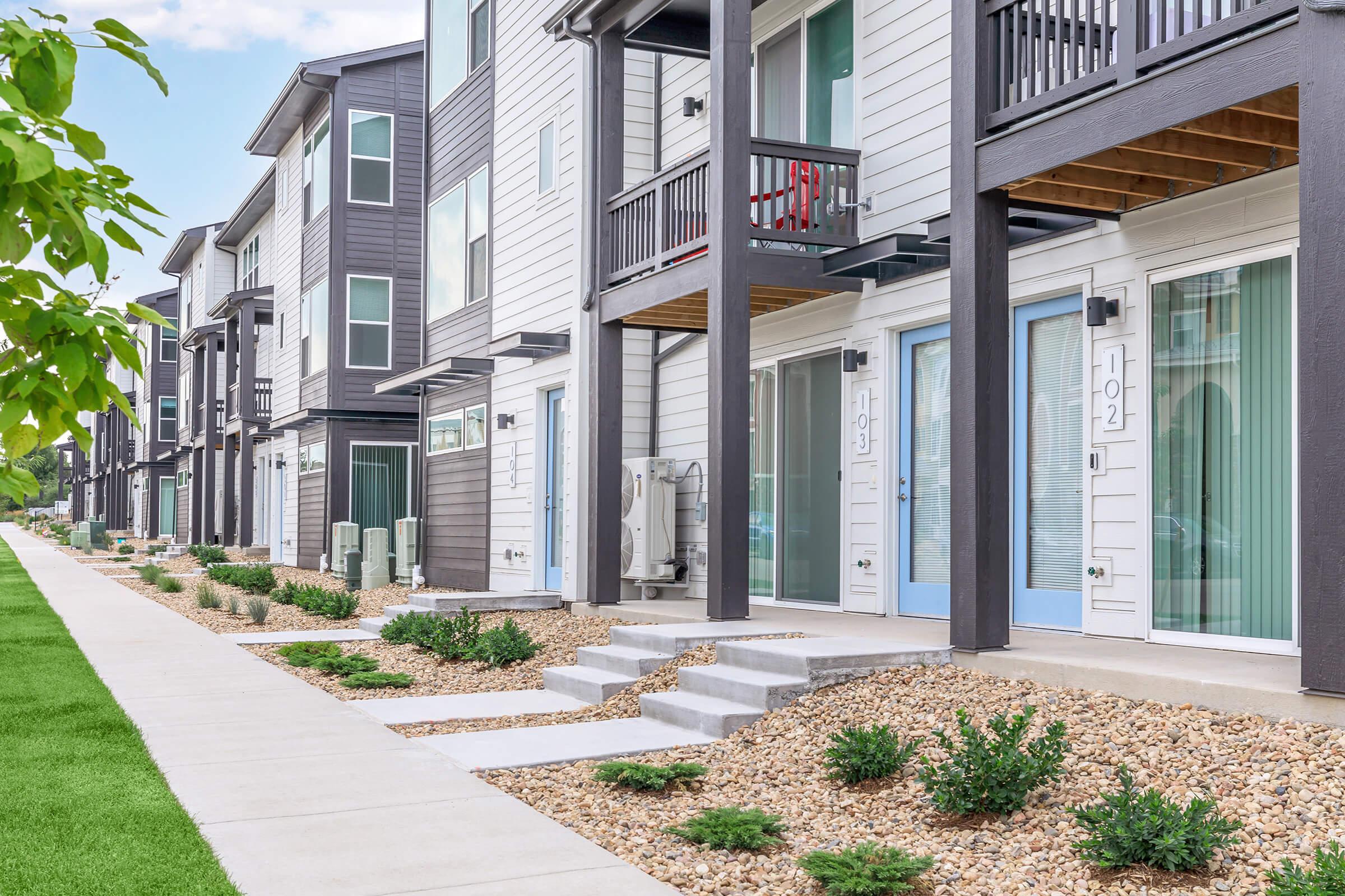 A modern row of multi-unit apartments, featuring light and dark colored exteriors, with small patios, stone landscaping, and a manicured lawn. The pathway leads up to the entrance doors numbered 102 and 103, with green shrubs lining the walkway.