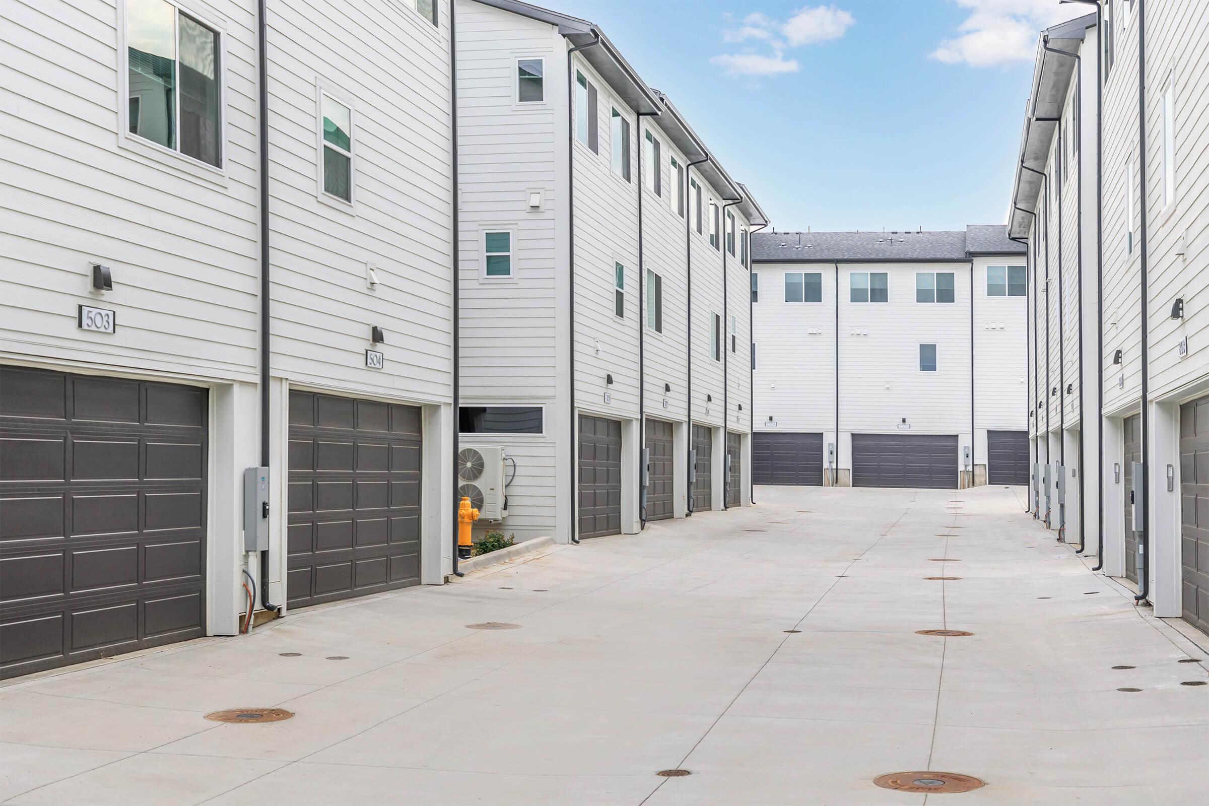 A view of a modern residential area featuring multiple two-story buildings with white siding. The image shows a paved alley lined with garages on either side, and clear blue skies above, creating a clean and organized appearance.