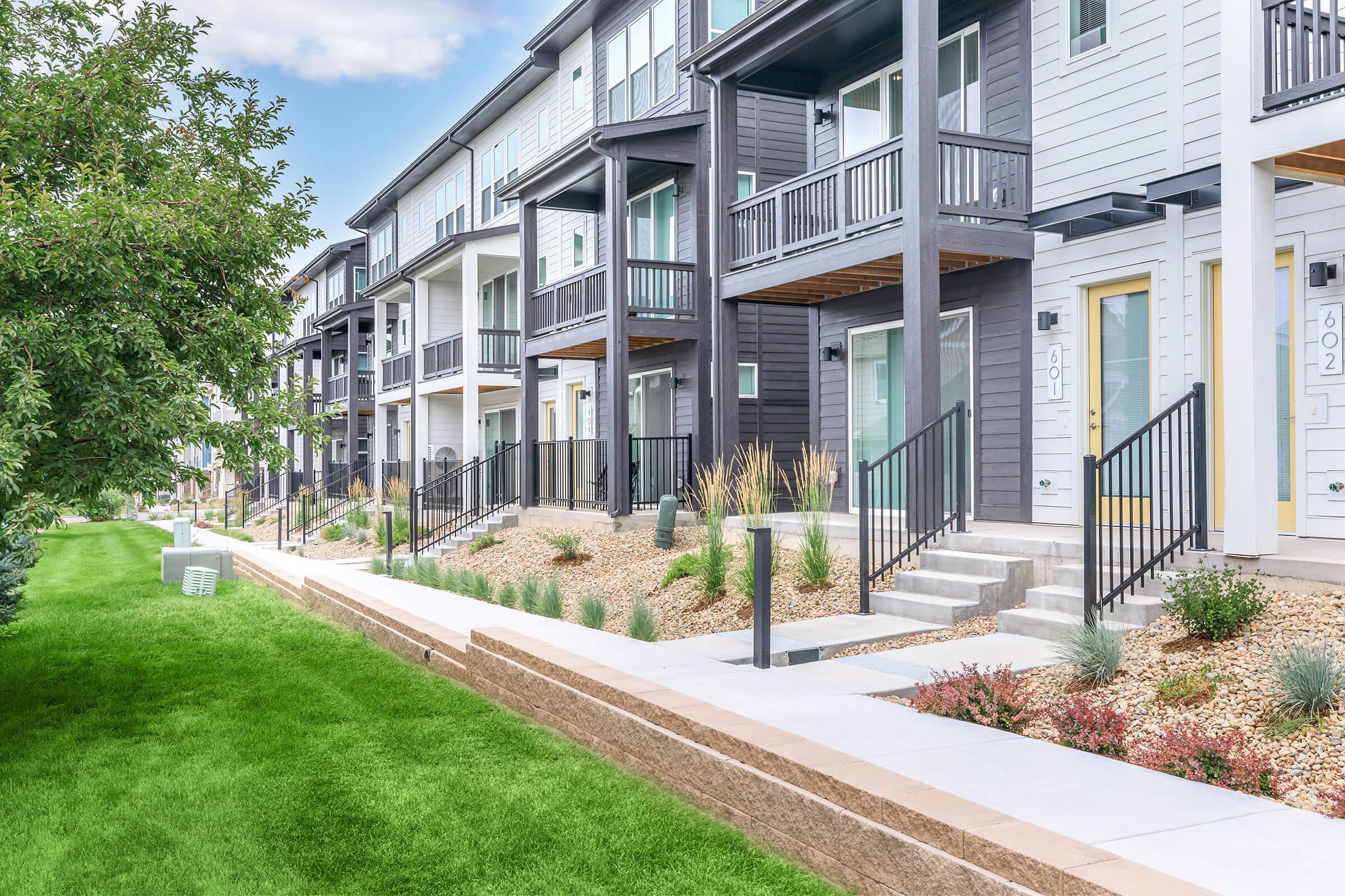 Row of modern townhouses with porches and balconies, featuring a variety of colors including dark gray and white. The landscape includes manicured lawns, planters with ornamental grasses, and a paved walkway. The setting is sunny, showcasing a well-maintained residential area.
