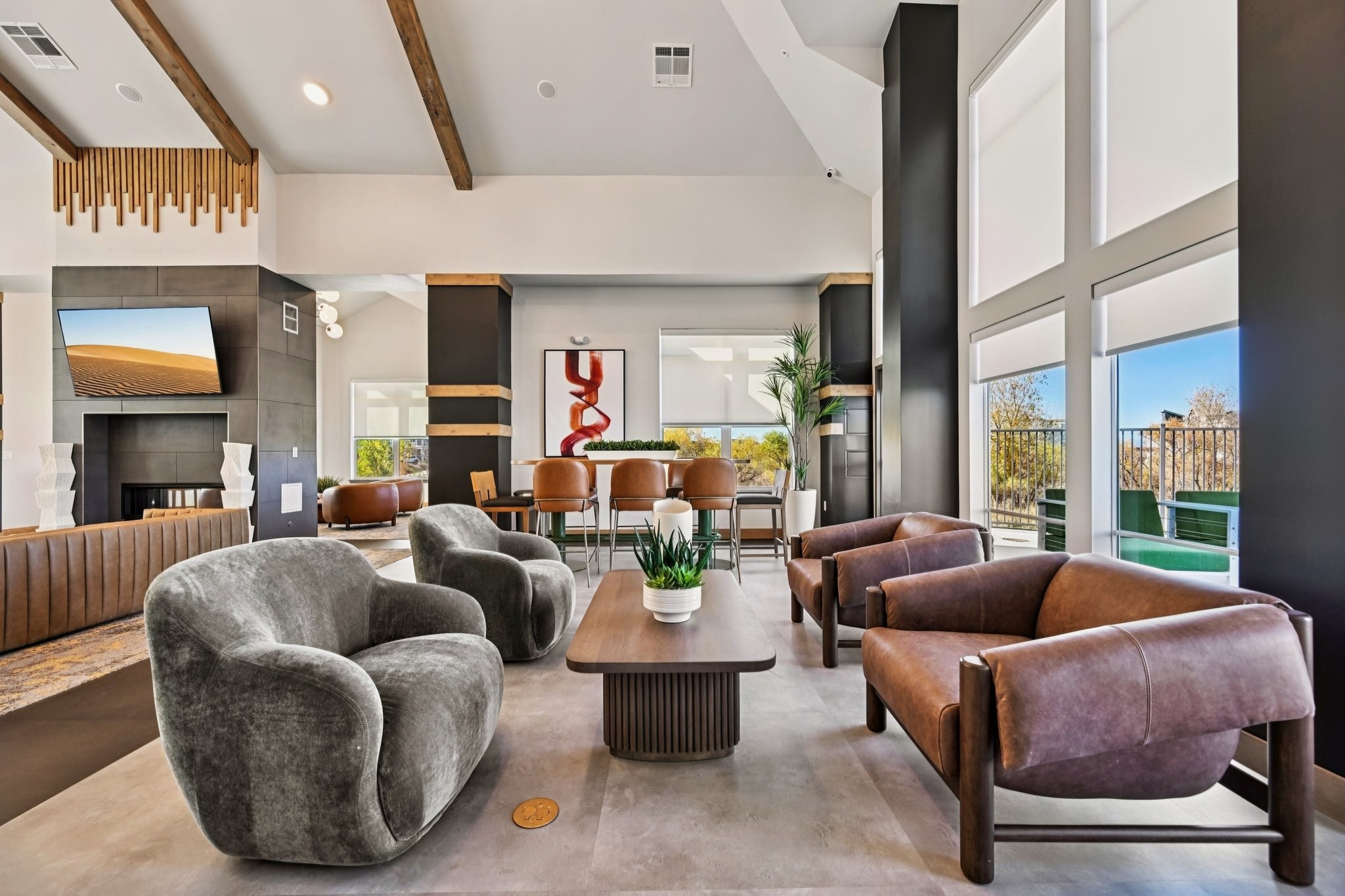 A modern living space featuring two plush grey chairs and two brown leather armchairs around a wooden coffee table. A large window offers natural light, and decorative plants add greenery. The walls are black with accents of wood and a red abstract art piece, creating a stylish and inviting atmosphere.
