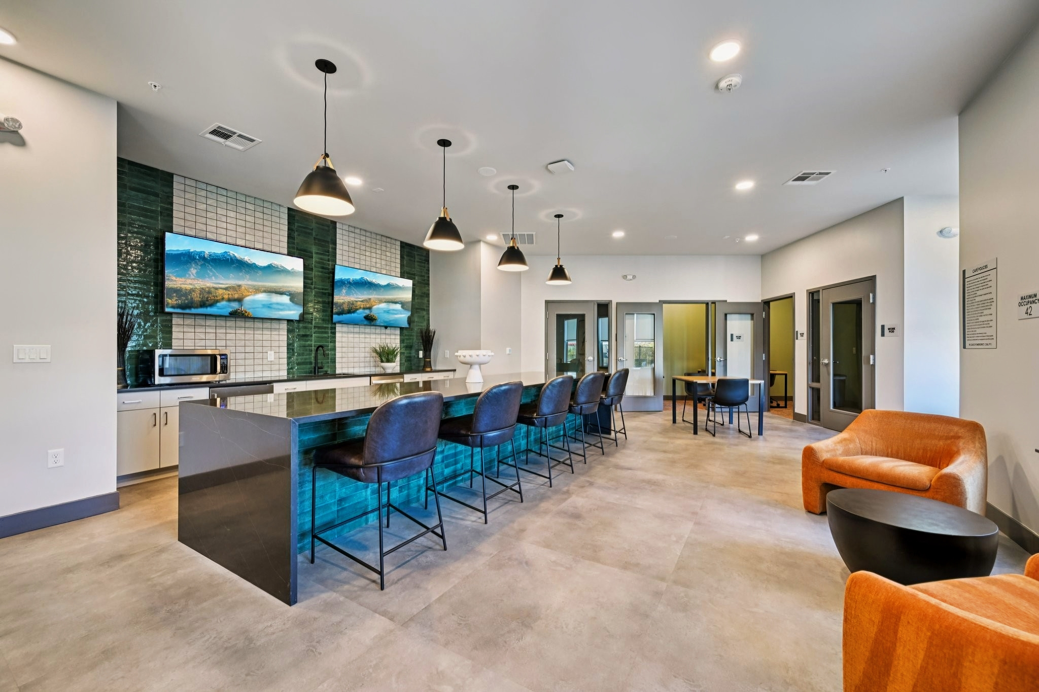 A modern common area featuring a kitchen island with barstools, a microwave, and two large-screen TVs displaying landscapes. The space has a contemporary design with pendant lighting, and includes a seating area with an orange armchair and black chair, along with neutral-colored walls and flooring.
