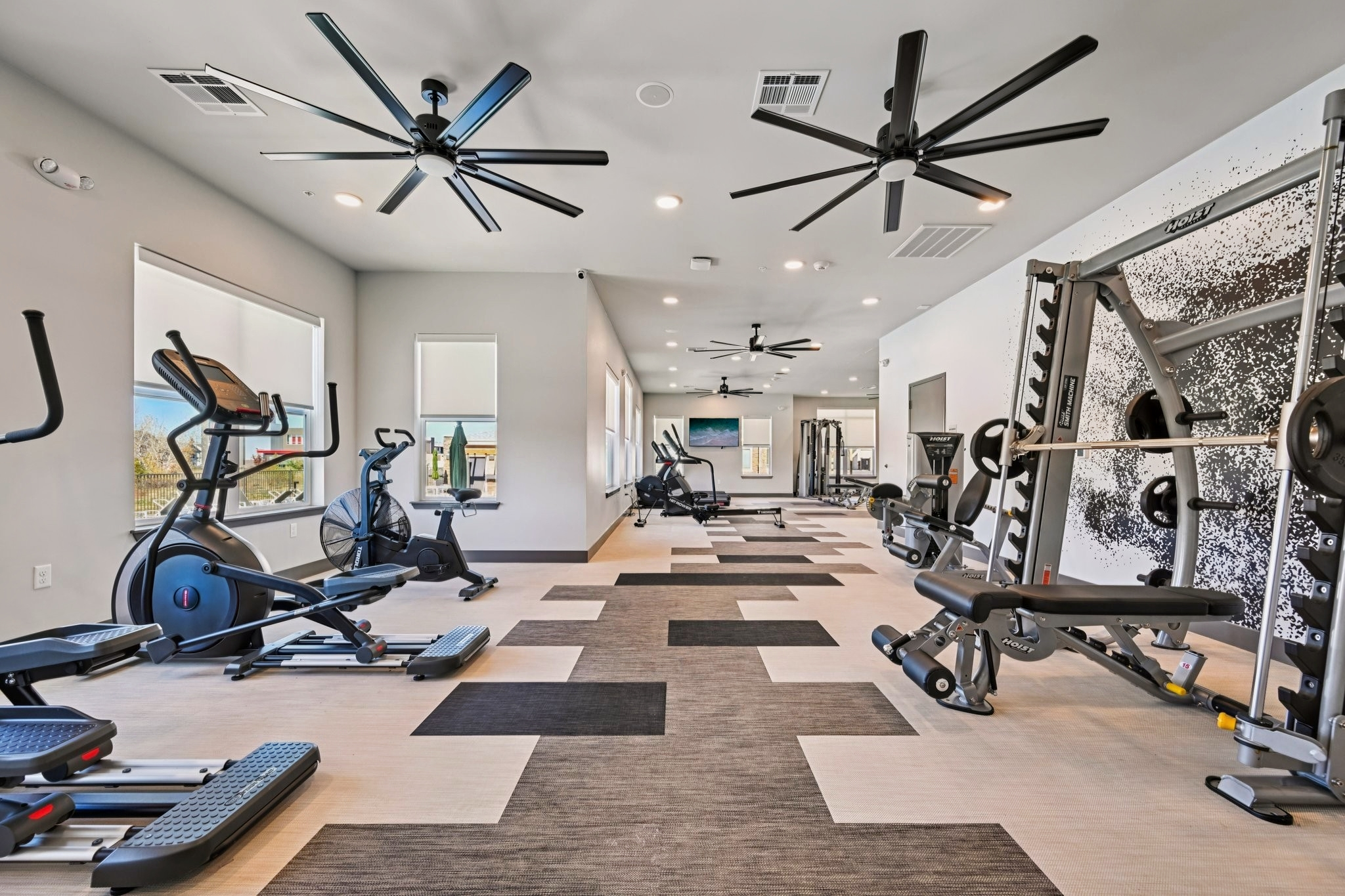 A modern gym interior featuring various exercise equipment including ellipticals, weight machines, and a free weights area. The space is well-lit with ceiling fans and large windows allowing natural light. The flooring has a mix of carpet and vinyl, creating a contemporary and inviting workout environment.