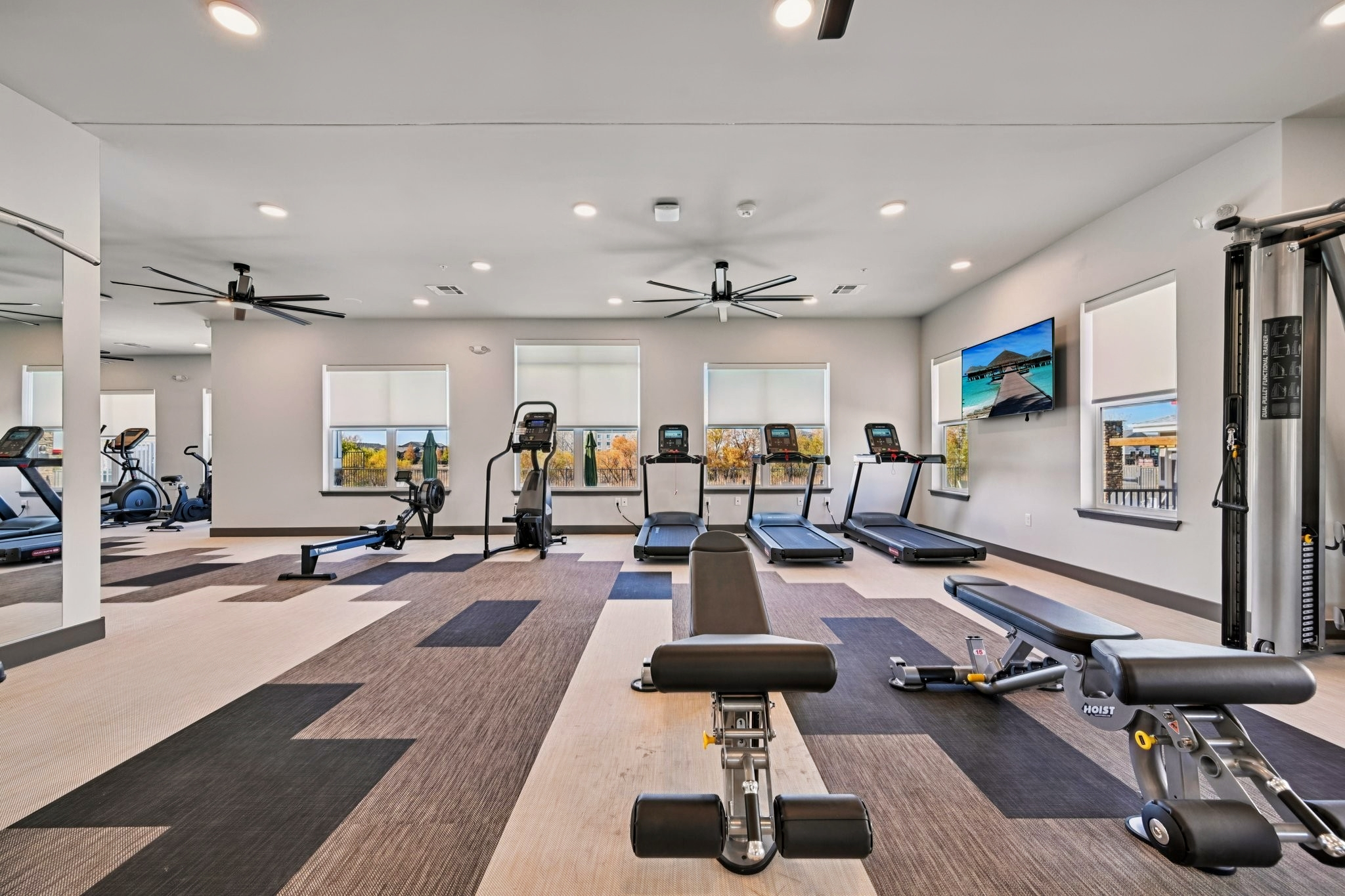 A modern gym interior featuring exercise equipment such as treadmills, a weight bench, and a multi-functional weight machine. Large windows provide natural light, and there are ceiling fans for ventilation. Mirrors line one wall, enhancing the spacious feel of the area.