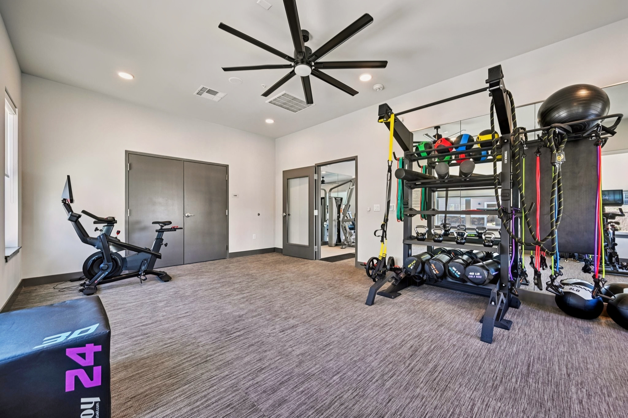 A modern gym interior featuring exercise equipment. There's a stationary bike, strength training station with resistance bands, and kettlebells neatly arranged. Large mirrors enhance the space, and a ceiling fan provides ventilation. The room is well-lit and designed for fitness activities.