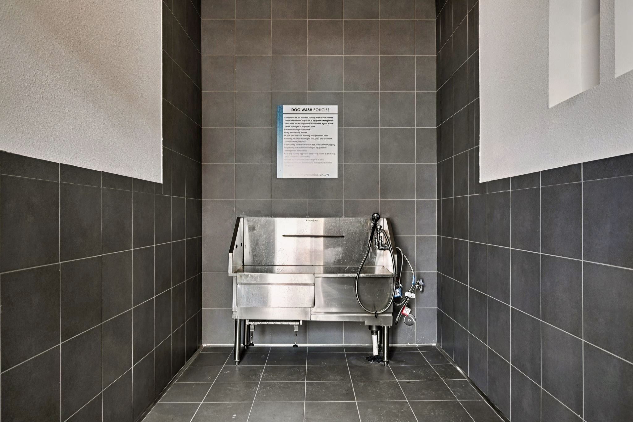 A stainless steel wash basin for dogs placed in a tiled room. The area has gray tiles on the floor and walls, and a sign with instructions or information is mounted on the wall above the wash basin. The space is clean and well-lit, designed for grooming pets.