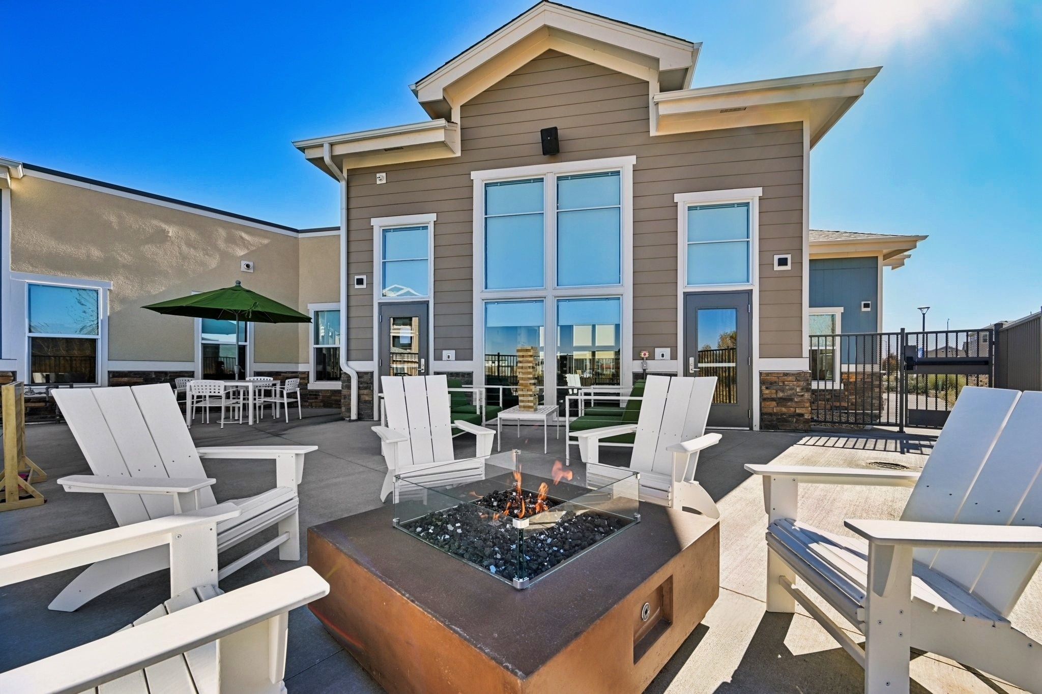 Outdoor seating area featuring white adirondack chairs surrounding a fire pit with glass stones. A green umbrella and patio tables are visible in the background, alongside large windows of a building under a clear blue sky. Sunlight casts a warm glow on the scene, creating an inviting atmosphere for relaxation.