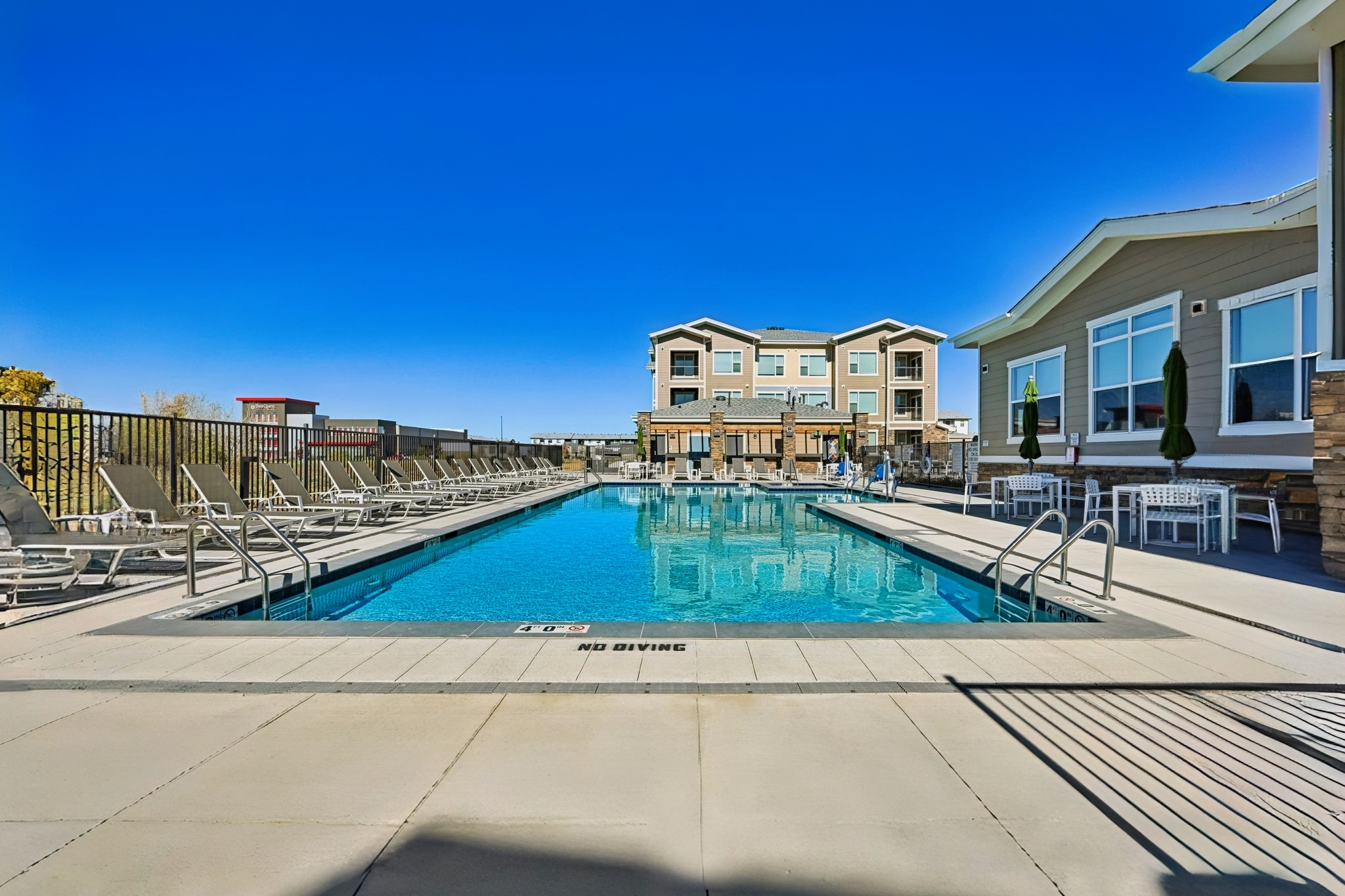 A clear blue swimming pool surrounded by lounge chairs, with a well-maintained deck. In the background, a modern building with multiple levels is visible. The sky is bright blue, creating a sunny and inviting atmosphere. The pool area appears tranquil and perfect for relaxation or recreation.