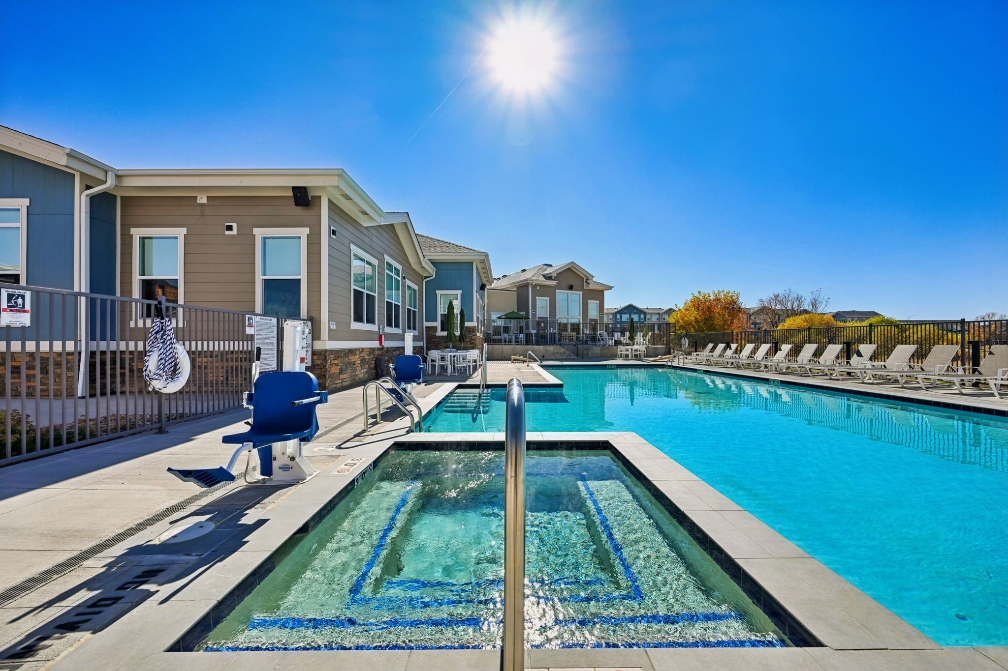 A clear blue swimming pool surrounded by lounge chairs, with a hot tub at the edge. In the background, a modern building with large windows and a sunny sky above.  The sun is shining brightly, casting reflections on the water's surface.
