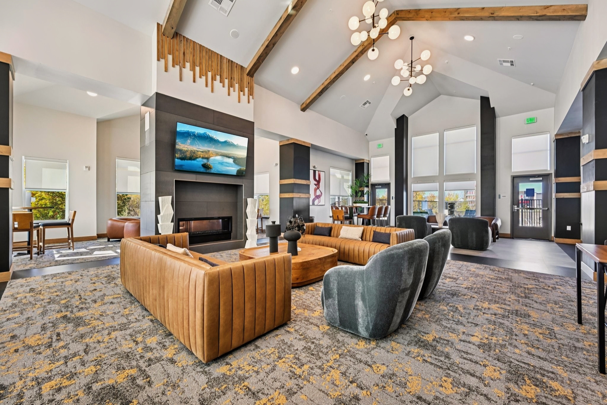 Modern interior of a spacious lounge area featuring high ceilings, large windows, and stylish furniture. The design includes a mix of patterned carpets, comfortable seating, a sleek fireplace, and a contemporary art piece on the wall. Natural light enhances the inviting atmosphere, creating a warm and welcoming space.