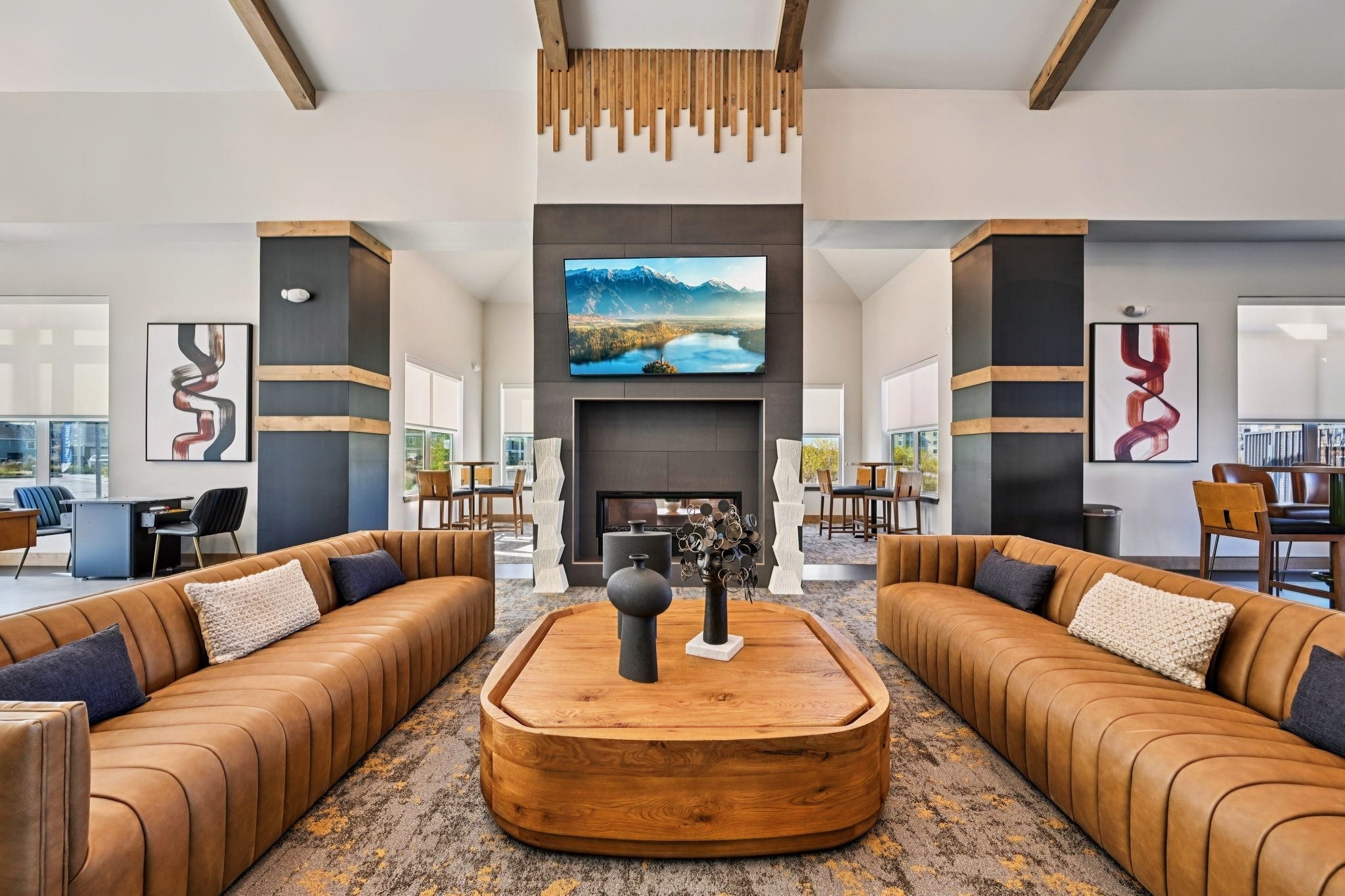Stylish living room featuring two brown leather sofas with decorative pillows, a large wooden coffee table, and a modern fireplace. A flat-screen TV displays a scenic mountain landscape, and the walls are adorned with contemporary artwork. The space has tall ceilings and large windows allowing natural light to filter in.