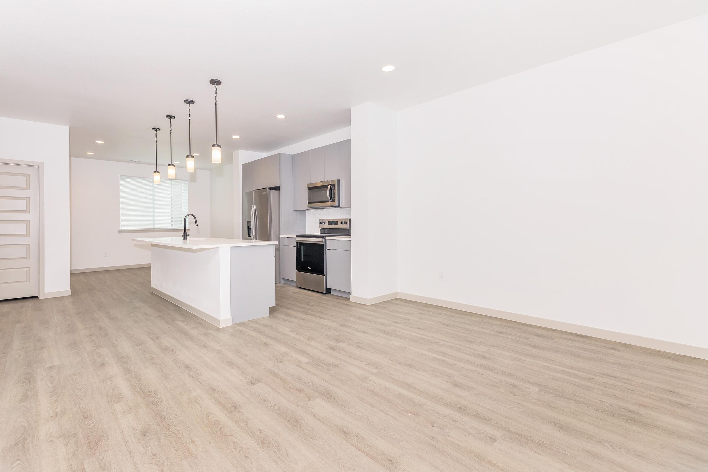 Bright, modern open-concept kitchen and living area featuring light wood flooring, stainless steel appliances, and a large island with pendant lighting. The space is accented by white walls, creating a clean and airy atmosphere, with a window providing natural light. Ideal for contemporary living.