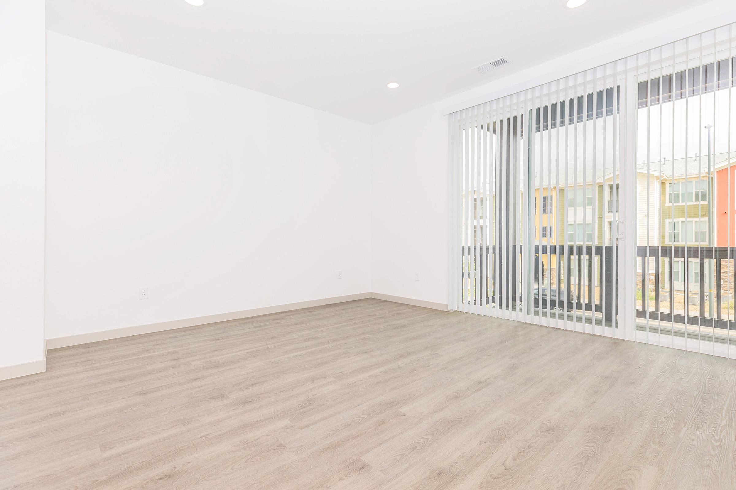Empty room with light-colored walls and laminate flooring. A large window with vertical white blinds lets in natural light, leading to a balcony view. The space is uncluttered and modern, suitable for various interior design styles.
