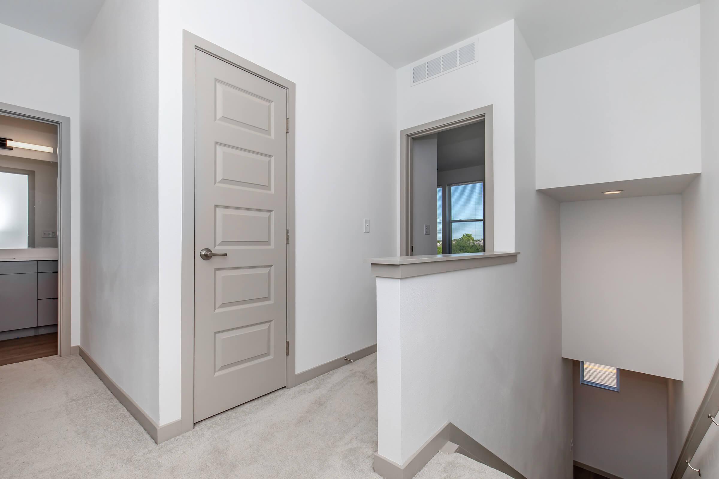 A spacious hallway with light-colored walls and carpet. There are two doors leading to rooms on the left, a window in an alcove on the right, and a staircase leading down at the far end. Natural light brightens the area, creating a welcoming atmosphere.