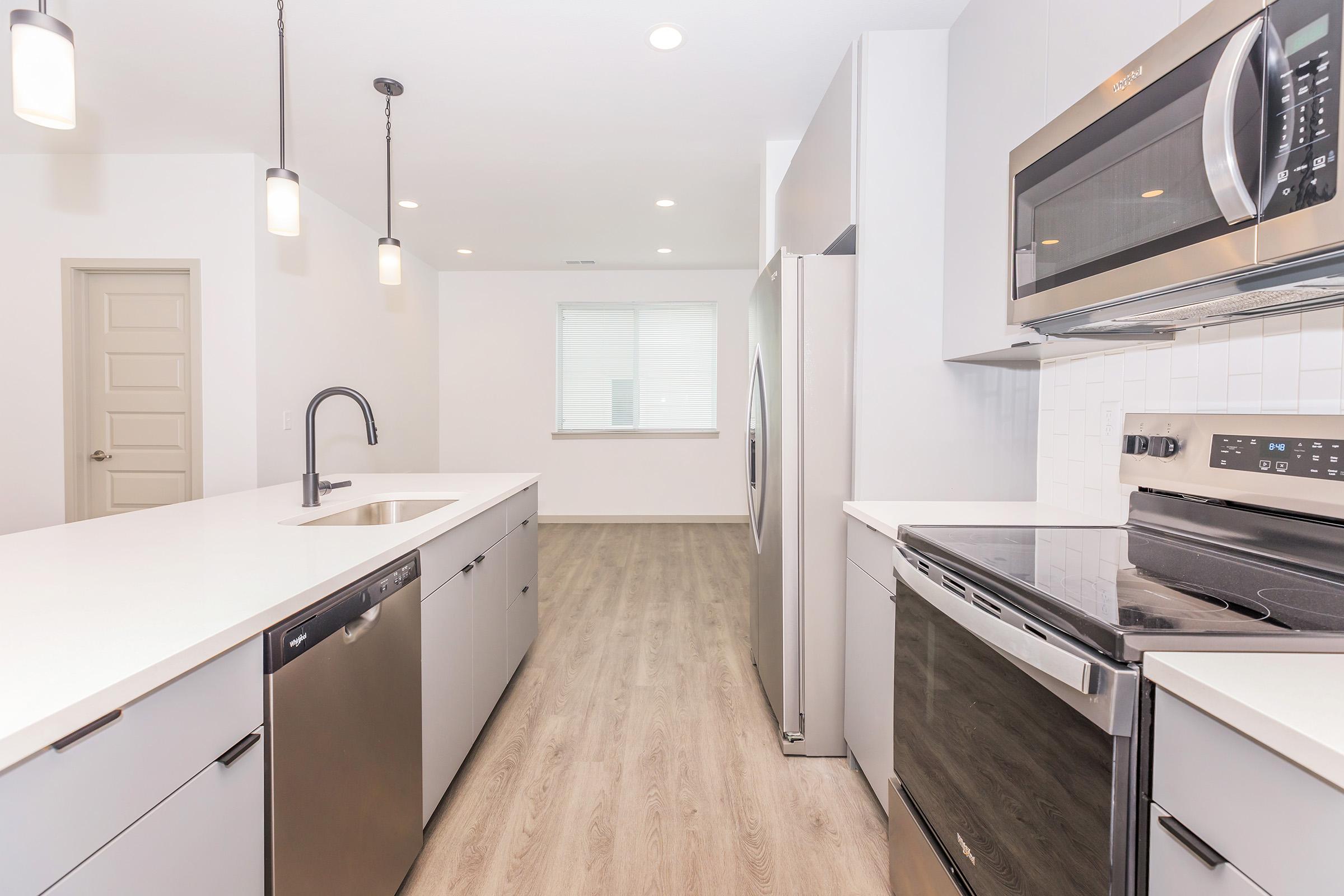 Modern kitchen featuring stainless steel appliances, including a refrigerator, microwave, and oven. The white cabinetry contrasts with the wooden floor, and there is a spacious island with a sink. Natural light filters in through a window, creating a bright and inviting atmosphere.