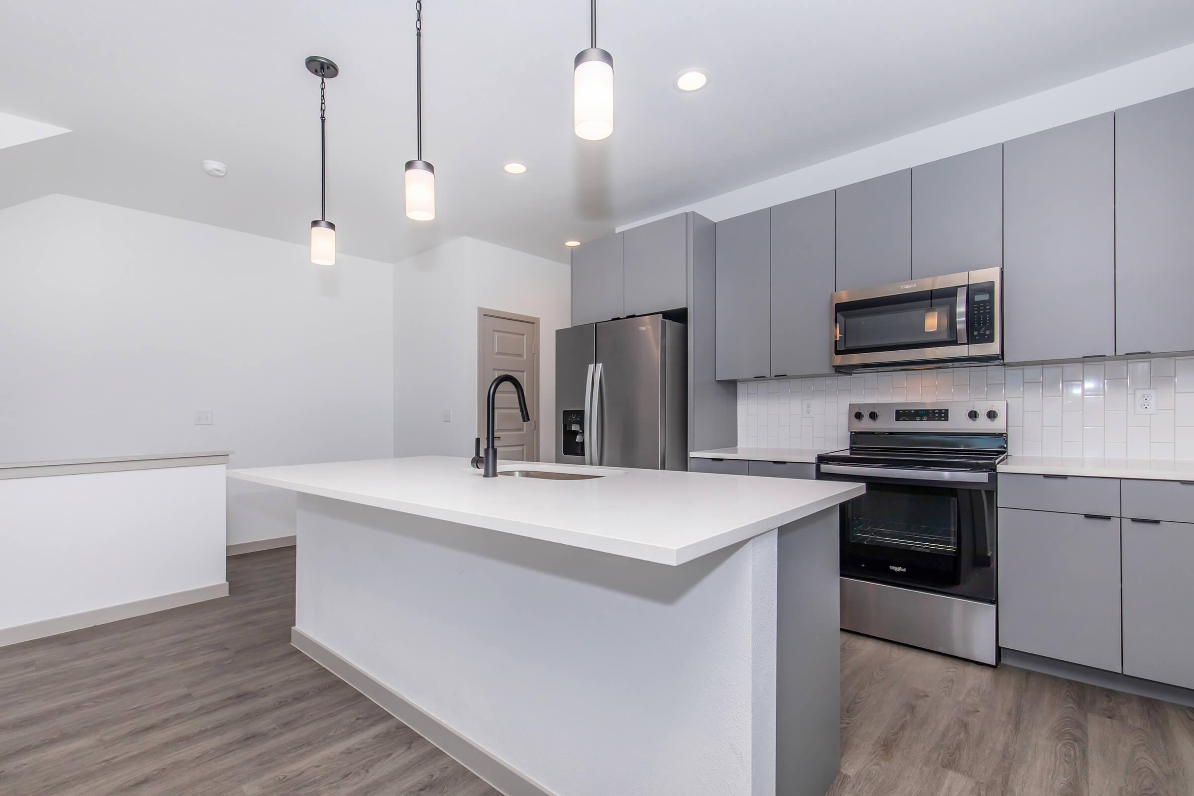 Modern kitchen featuring gray cabinets, stainless steel appliances including an oven and microwave, a large island with a sink, and pendant lighting. The flooring is a light-colored laminate, and the overall design is sleek and contemporary, with an open layout leading to another room.