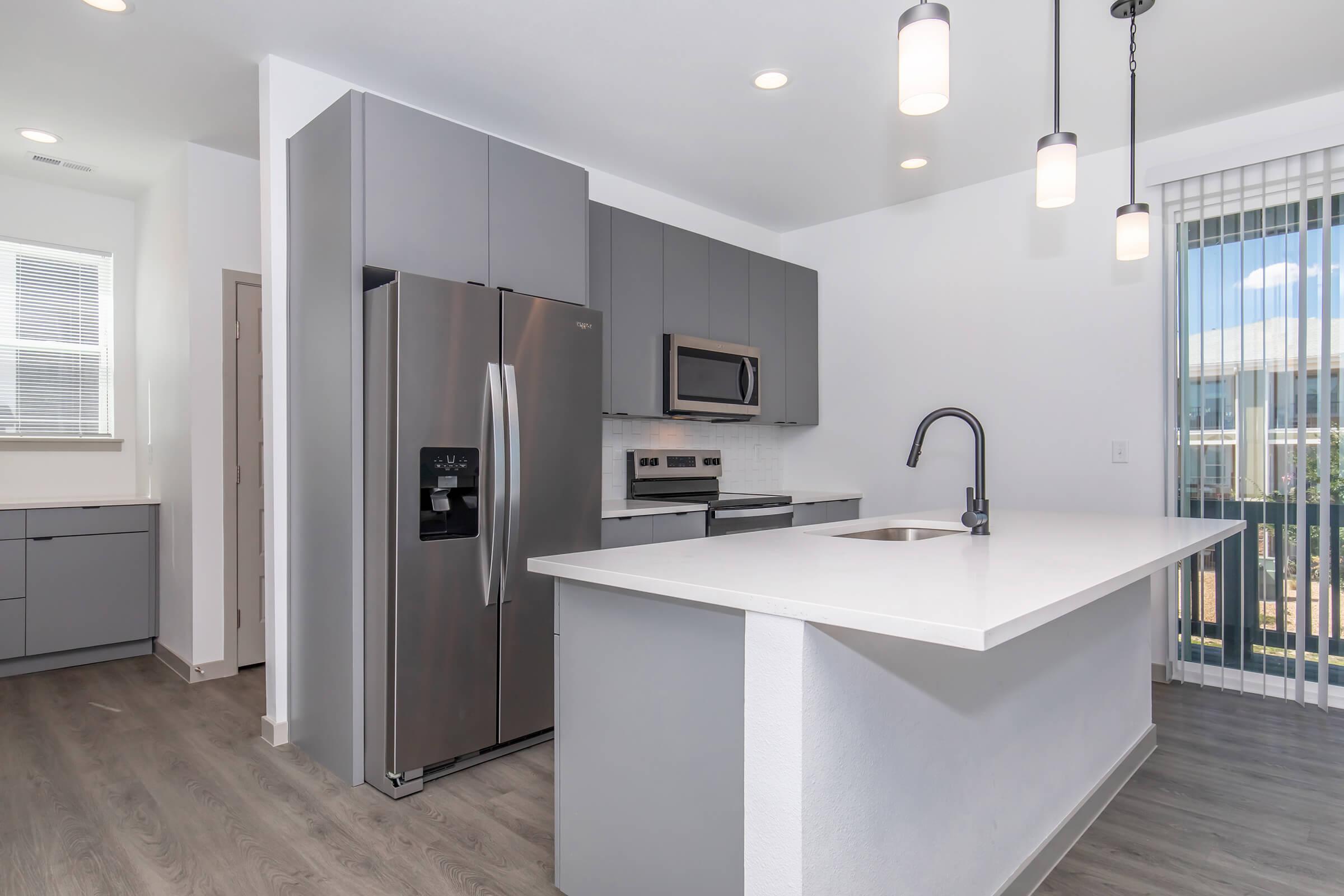 Modern kitchen featuring sleek gray cabinets and appliances, including a stainless steel refrigerator and microwave. A white countertop with a single basin sink, pendant lighting overhead, and a large window providing natural light. The flooring is a light wood finish.