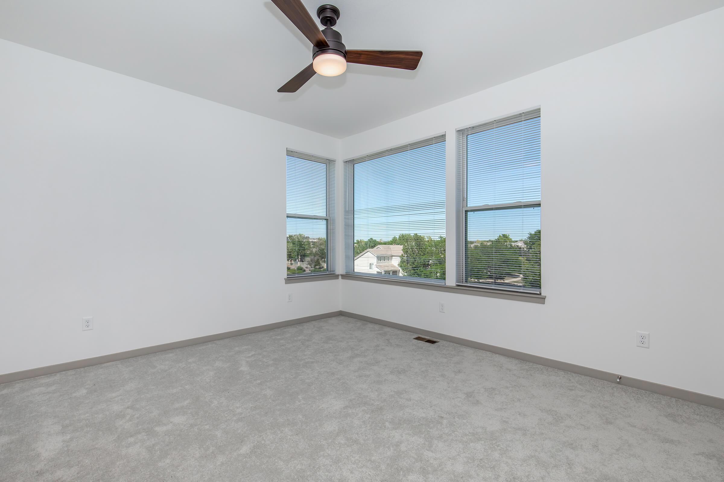 Empty room with light gray carpet, featuring large windows allowing natural light in. A ceiling fan with a light fixture is installed. The walls are painted white, and the room has a minimalistic design, creating a spacious and airy feel.