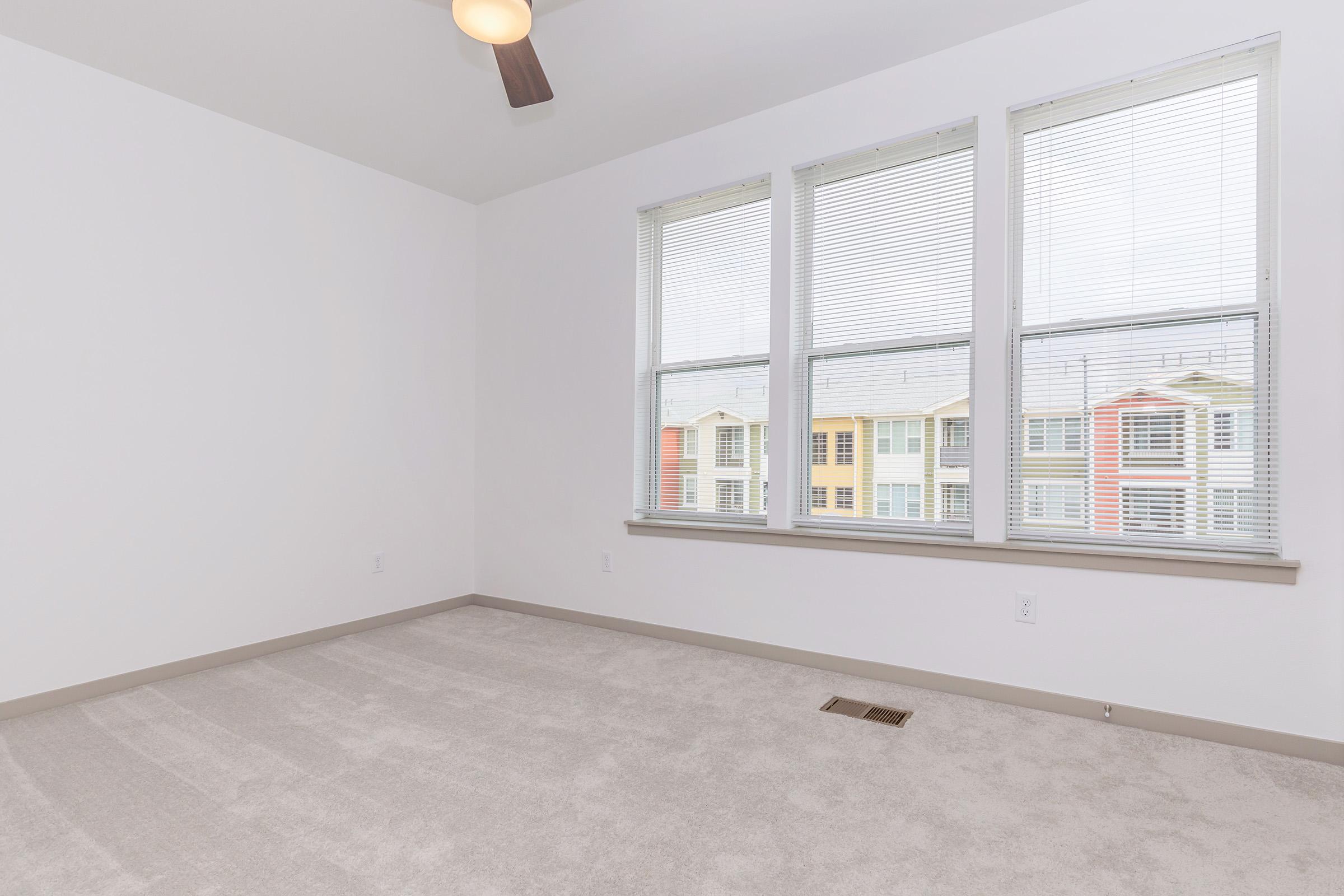 A spacious, empty room with light-colored walls and a ceiling fan. The floor is covered in beige carpet. Three large windows with blinds allow natural light, revealing a colorful view of buildings outside. The overall atmosphere is bright and minimalist, suitable for various interior design options.