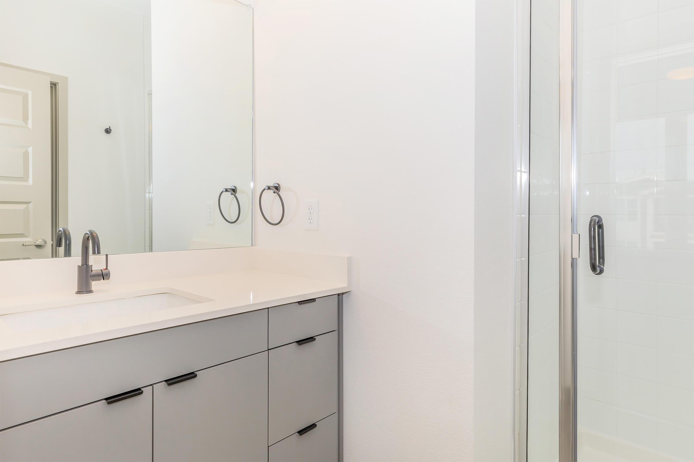 A modern bathroom featuring a gray vanity with a white countertop, two round mirror above the sink, and a shower with a clear glass door. The walls are painted white, and the floor is tiled, creating a clean and contemporary look. The design emphasizes simplicity and functionality.