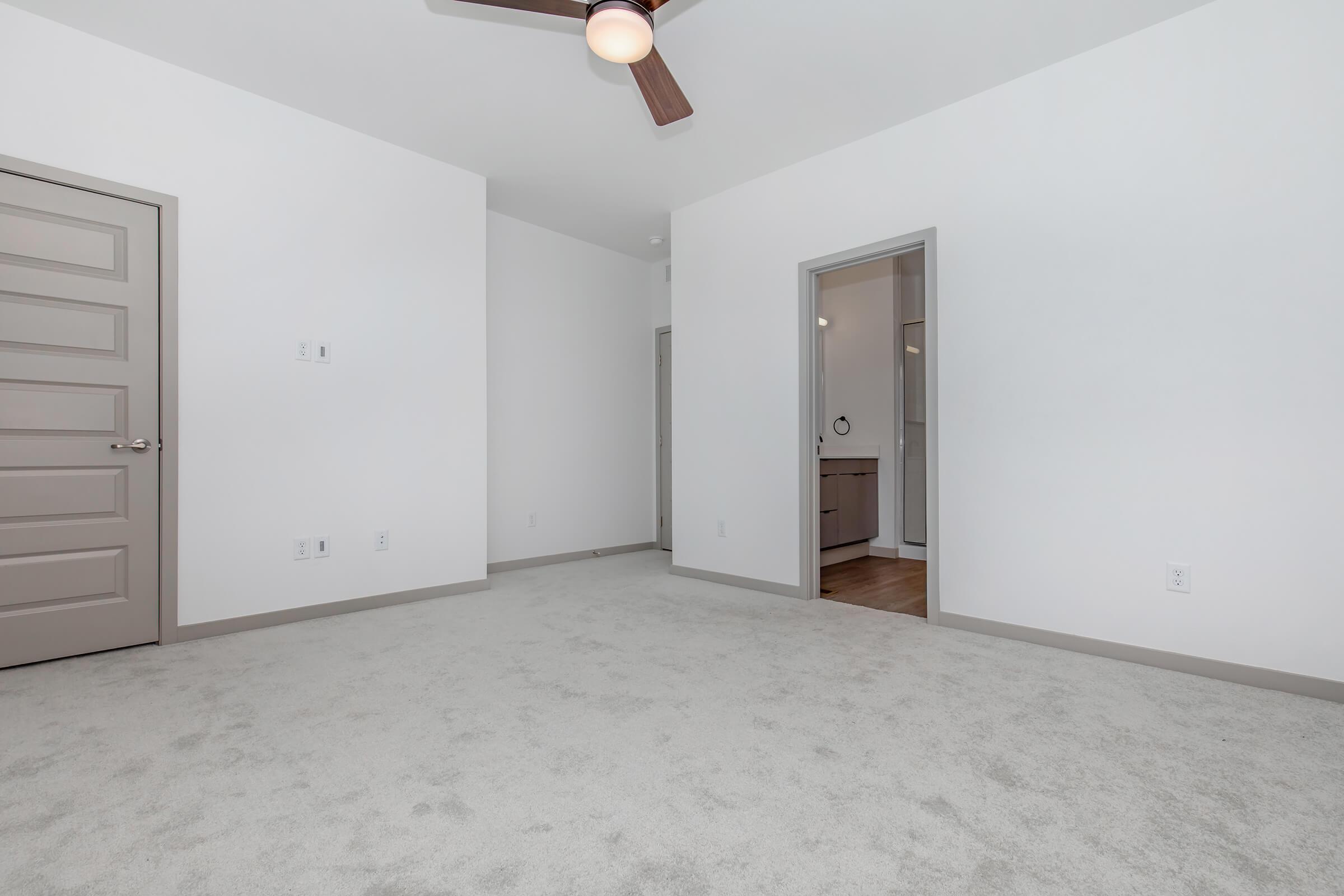 A spacious, empty room with light gray walls and carpet. A ceiling fan is mounted above, and there's a closed door on the left. An open doorway leads to a bathroom area in the background, which has a simple, modern design. The room is well-lit with ample natural light.